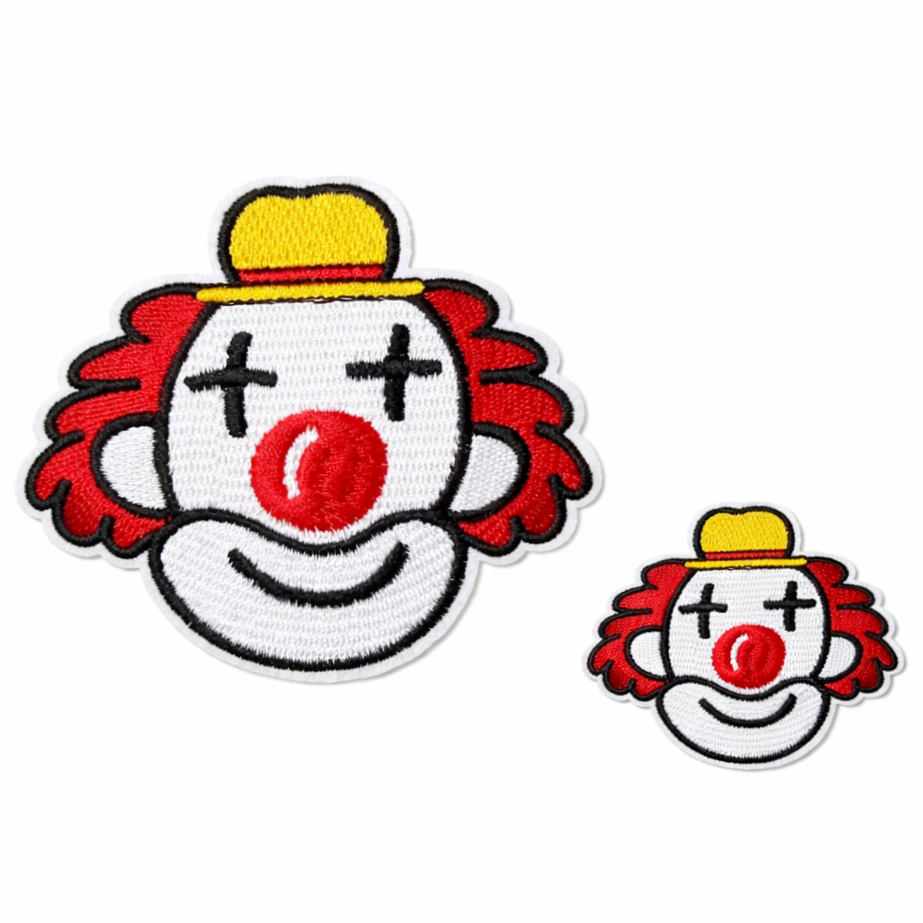 Gestickter Karneval-Aufnäher in Form eines Clownkopfes in zwei Größen auf weißem Hintergrund. Der Clown hat rote, lockige Haare, eine große runde rote Nase, ein freundlich lächelndes Gesicht und schwarze Augen in Kreuzform. Er trägt einen gelben Hut mit rotem Band. Die Stickerei ist in den Farben Rot, Weiß, Gelb und Schwarz gehalten und besitzt eine klare, kontrastreiche Umrandung. Links ist der Aufnäher in größerer Ausführung zu sehen, rechts daneben die kleinere Variante