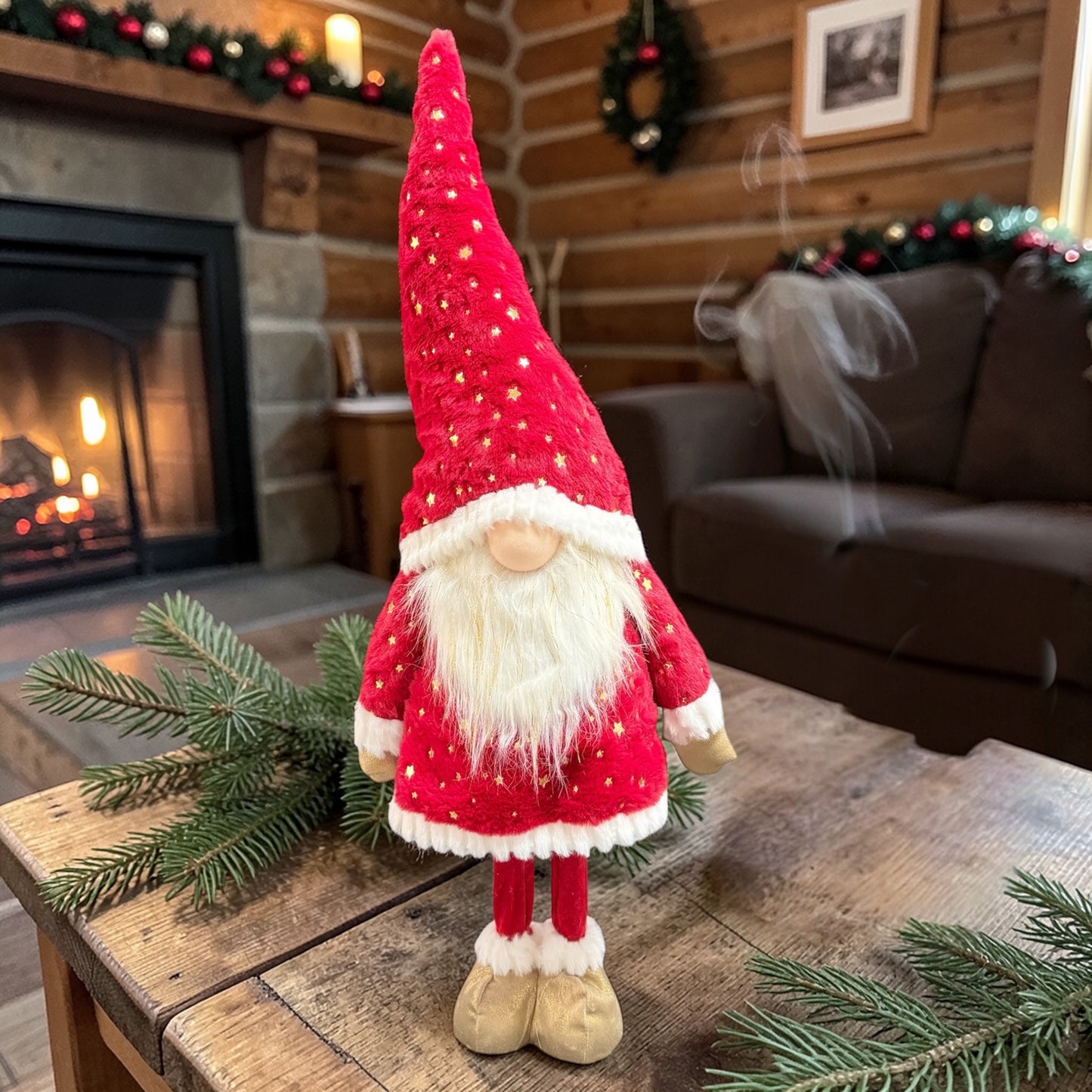 Rote Wichtelfigur aus Stoff mit weißem Bart auf einem Tisch mit Tannenzweigen vor einem Kamin und weihnachtlicher Dekoration.