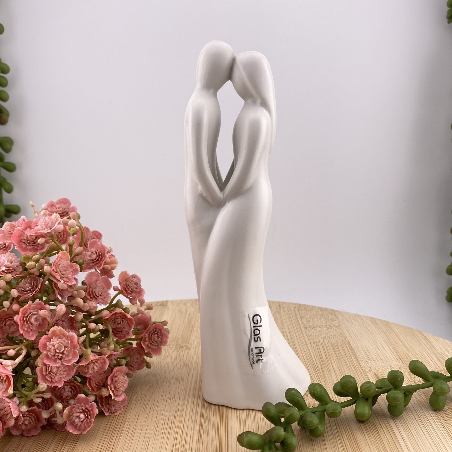 Eine weiße Dekofigur in Form eines verliebten Paares. Das Ehepaar fasst einander an den Händen und hat die Köpfe aneinander gelehnt. Im Ambiente platziert.