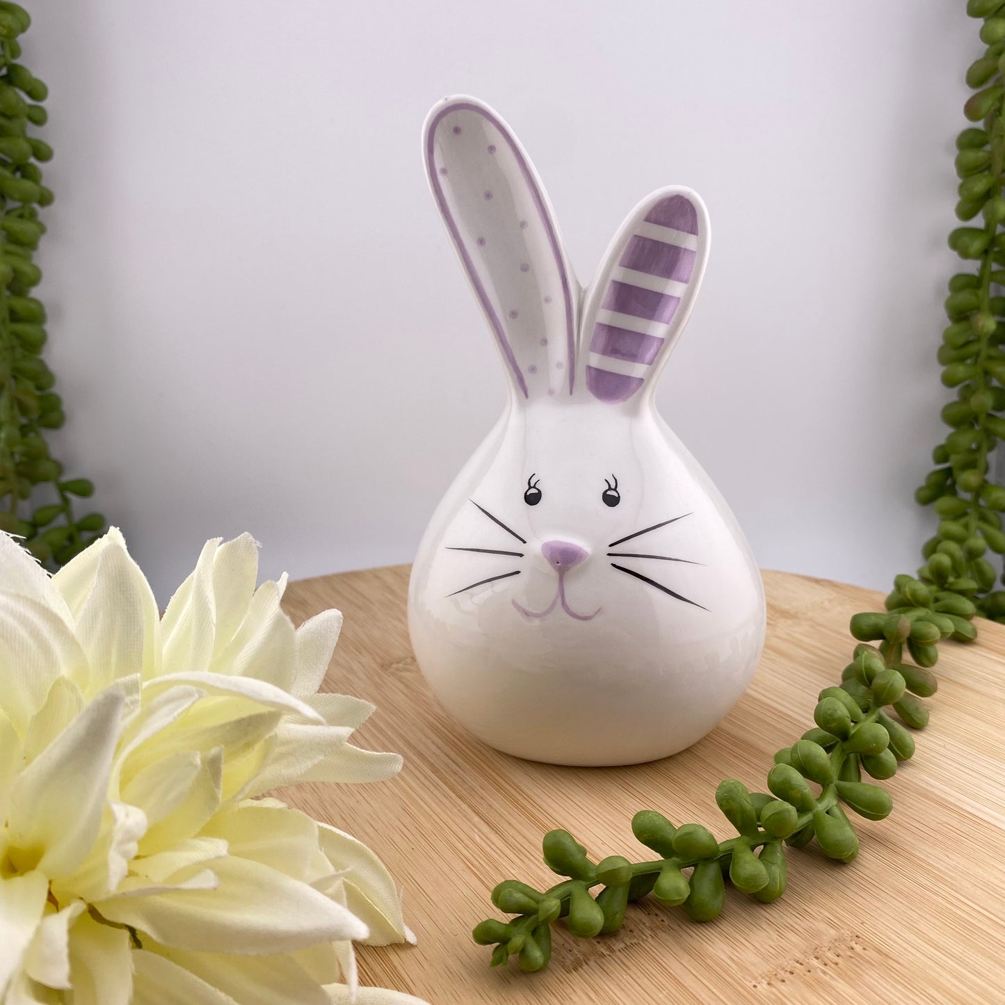 Ein weißer Keramik Deko Osterhase in Kugelform lächelt zufrieden und hat die Ohren gespitzt. Er hat lilane Akzente und ist Ambiente platziert.