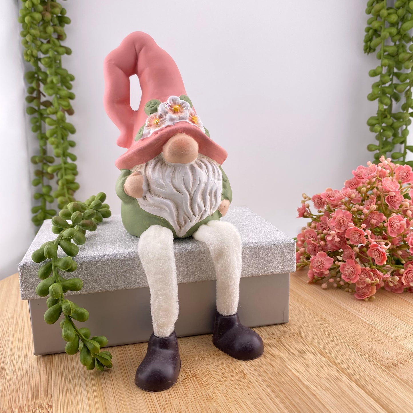 Eine Dekofigur in Form eines Wichtels bzw. Gnoms, der gemütlich sitzt. Das Dekoobjekt hat eine rosane Zipfelmütze auf, trägt einen langen weißen Bart und ein grünes Oberteil. Der Dekozwerg ist im Ambiente platziert.