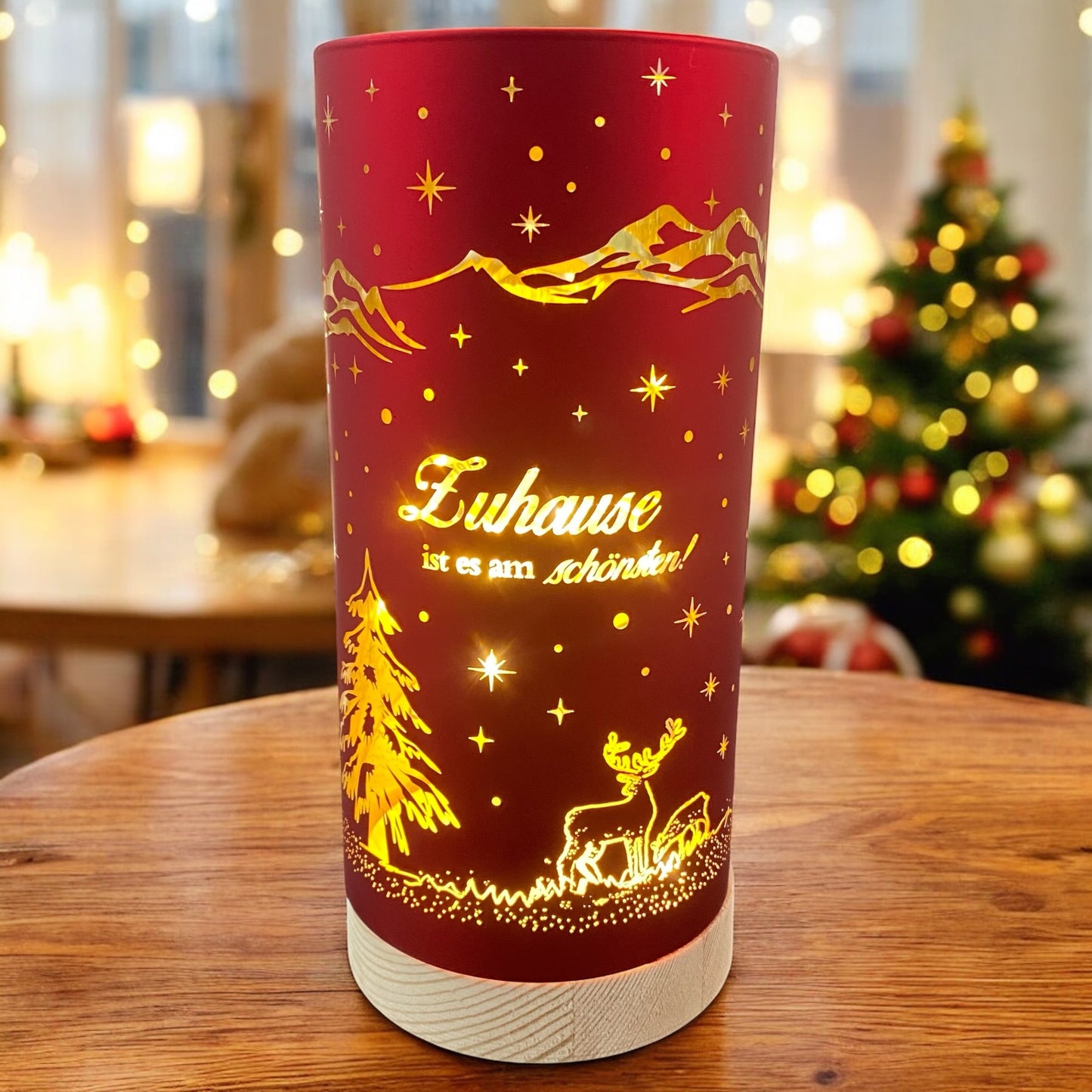 Großes rotes Deko-Licht mit goldener Aufschrift „Zuhause ist es am schönsten!“ und leuchtendem Hirschmotiv, auf Holztisch vor Weihnachtsbaum.