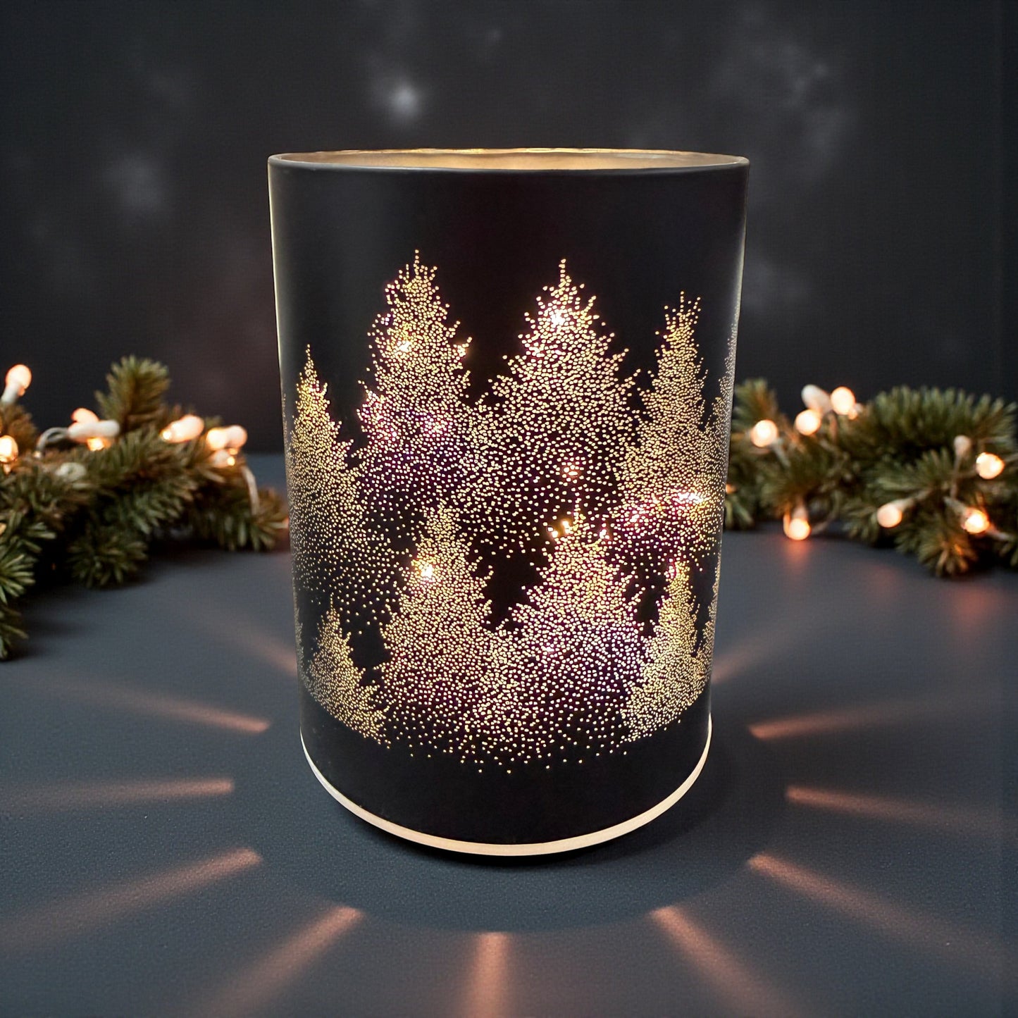 Ein zylindrisches schwarzes Dekolicht mit perforiertem Tannenbaum-Motiv und hellem Innenlicht steht auf einer Oberfläche vor Tannenzweigen und einer Lichterkette.