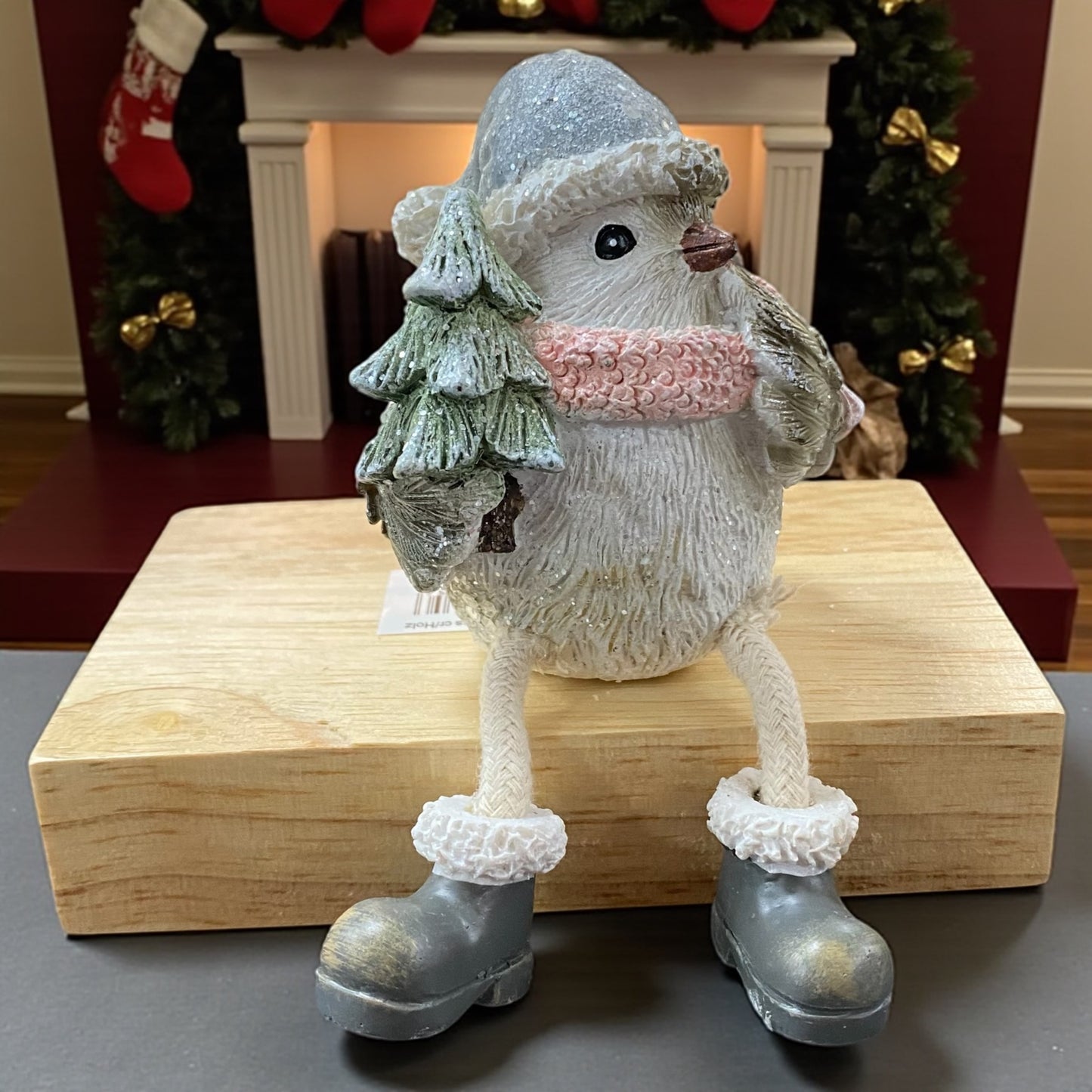 Winterliche Vogel-Figur mit Mütze und rosa Schal, die einen kleinen Tannenbaum hält, auf einem Holzblock vor festlicher Kulisse.