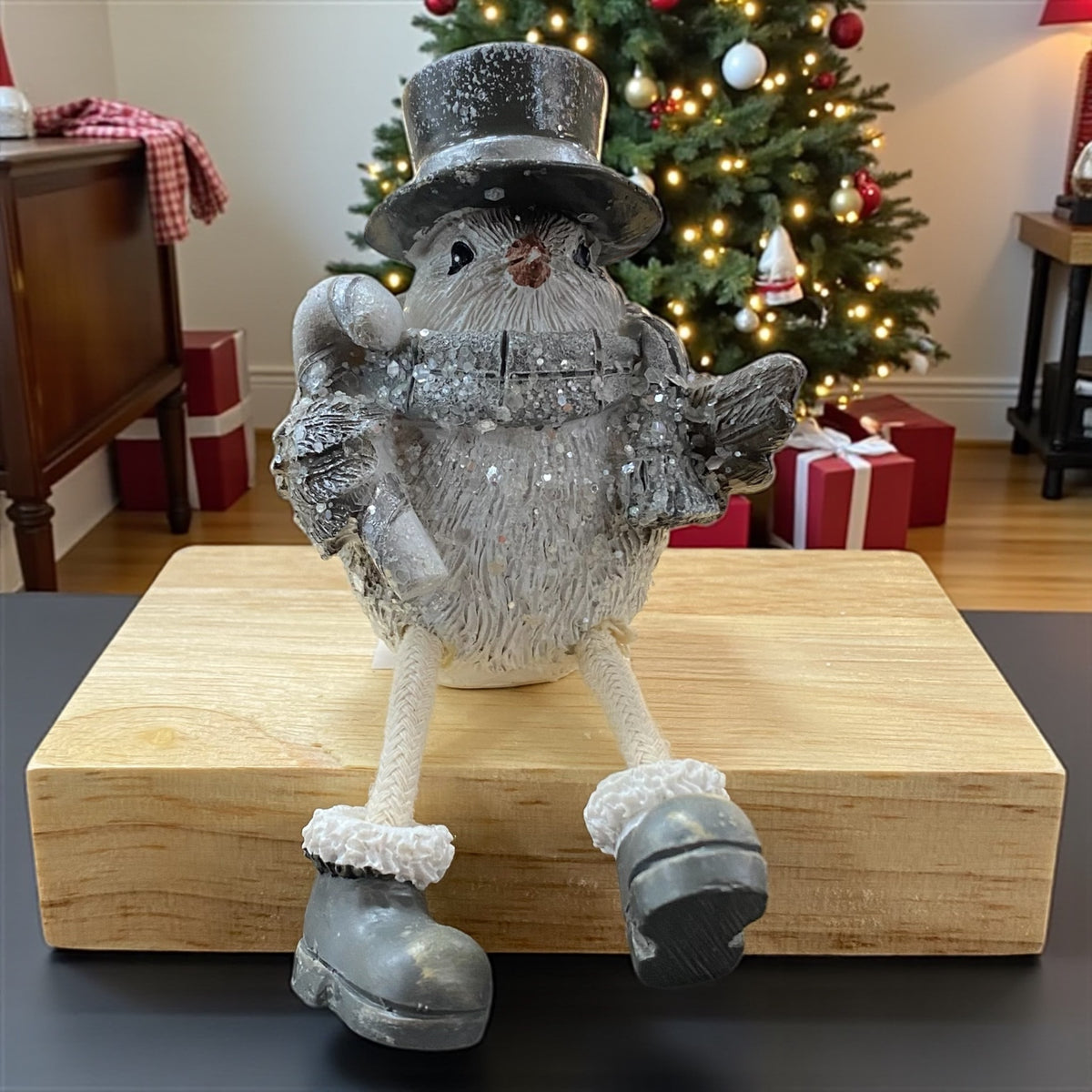 Winterliche Vogel-Figur mit Zuckerstange und Zylinderhut als Kantenhocker auf einem Holzblock vor Weihnachtsbaum und Geschenken.