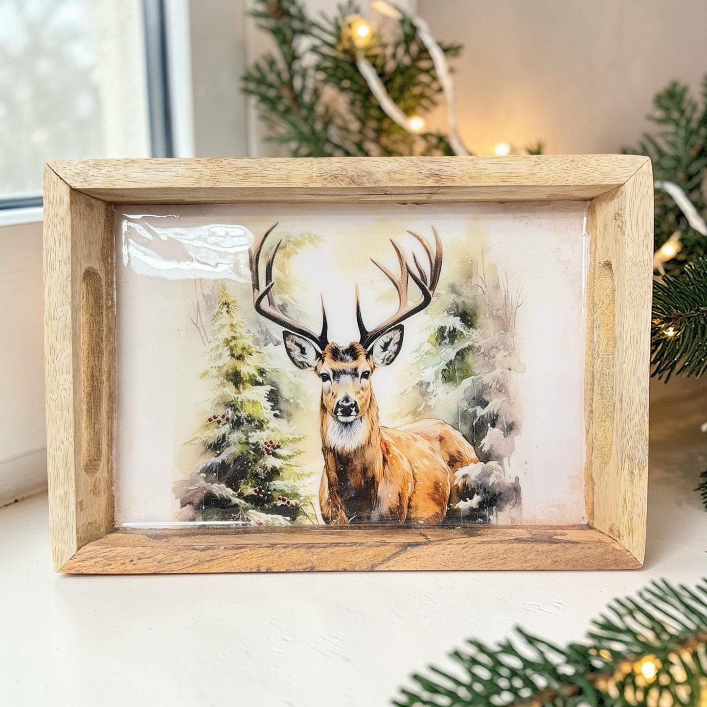 Rechteckiges Tablett mit Holzrahmen und Hirschmotiv auf einer Fensterbank platziert. Umgebung mit Tannenzweigen und Lichterkette, weihnachtliche Dekoration im Hintergrund.