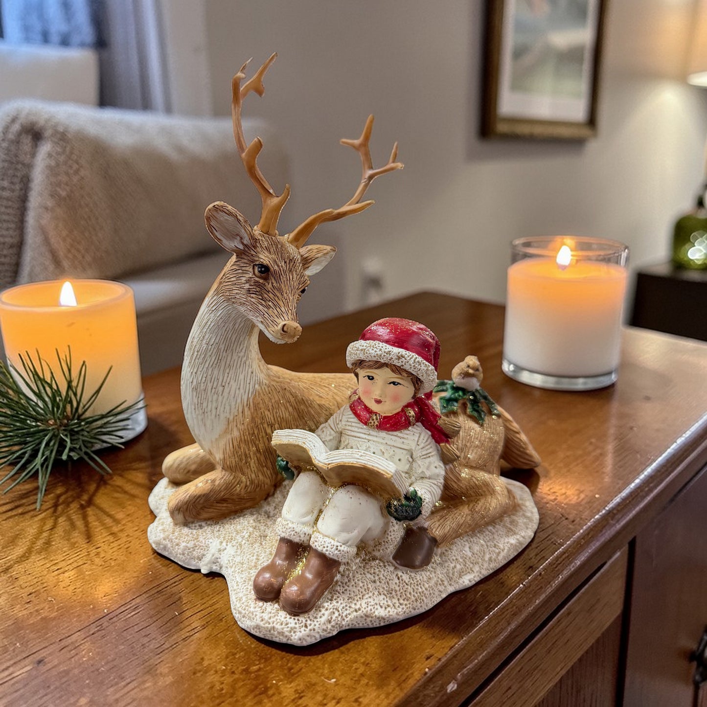 Eine dekorative Figur mit einem sitzenden Kind, das ein Buch liest, ist zusammen mit einem Hirsch und einem Reh auf einer Holzoberfläche neben Kerzen platziert.