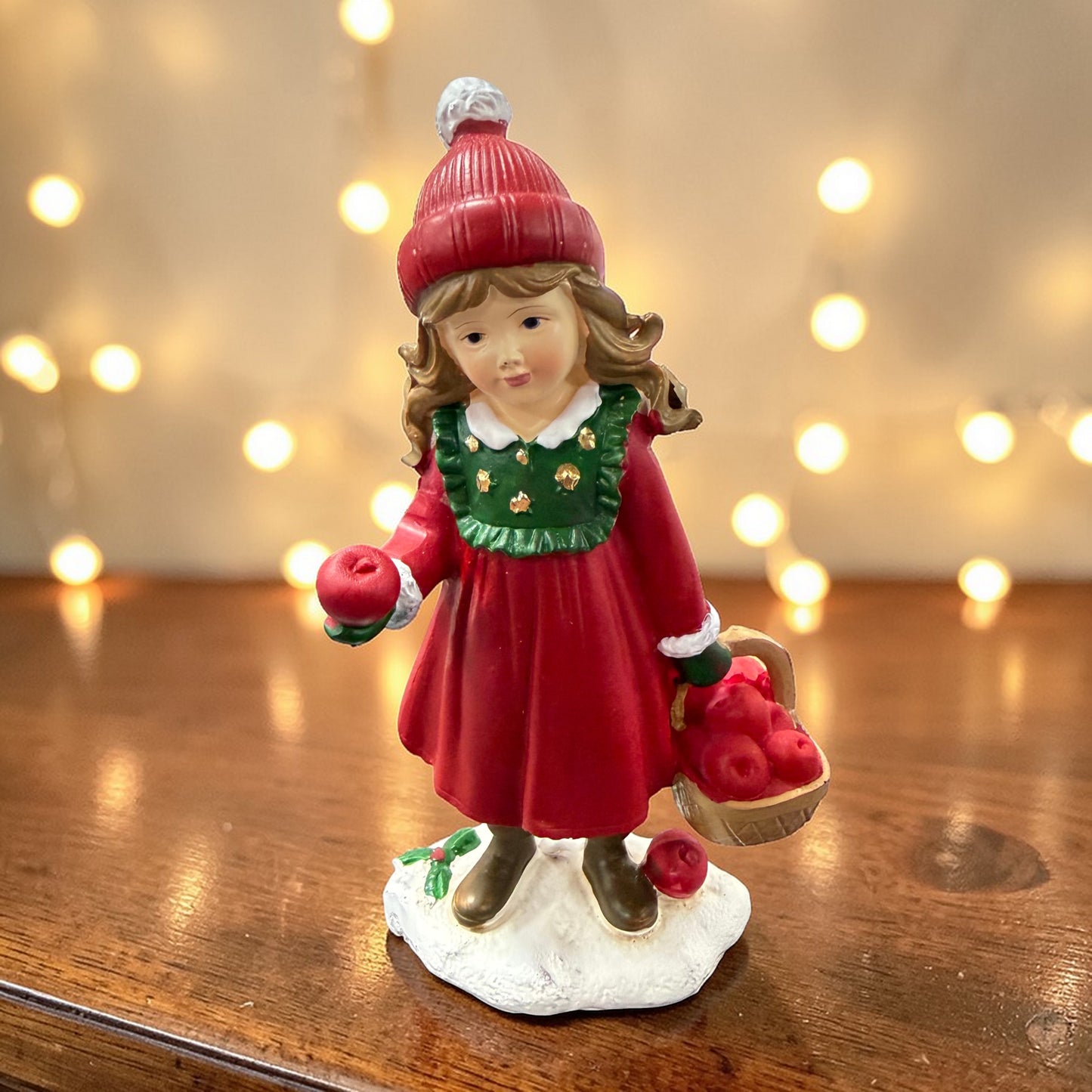 Eine stehende Mädchenfigur mit roter Mütze hält einen Apfel und trägt einen Korb mit weiteren Früchten.