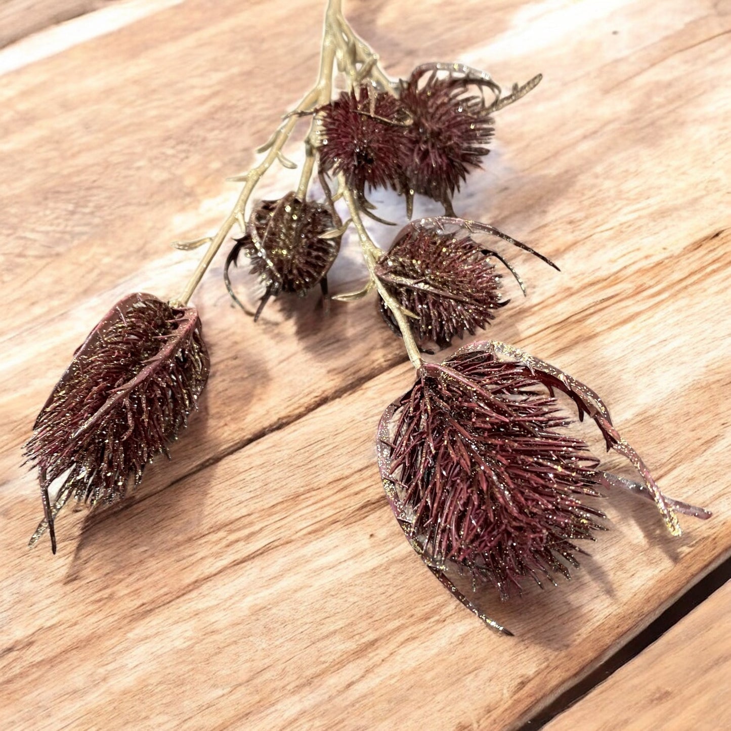 Künstlicher Distelzweig in Bordeaux und Gold mit mehreren glitzernden Blütenköpfen und feinen, verzweigten Stielen auf hölzernem Hintergrund