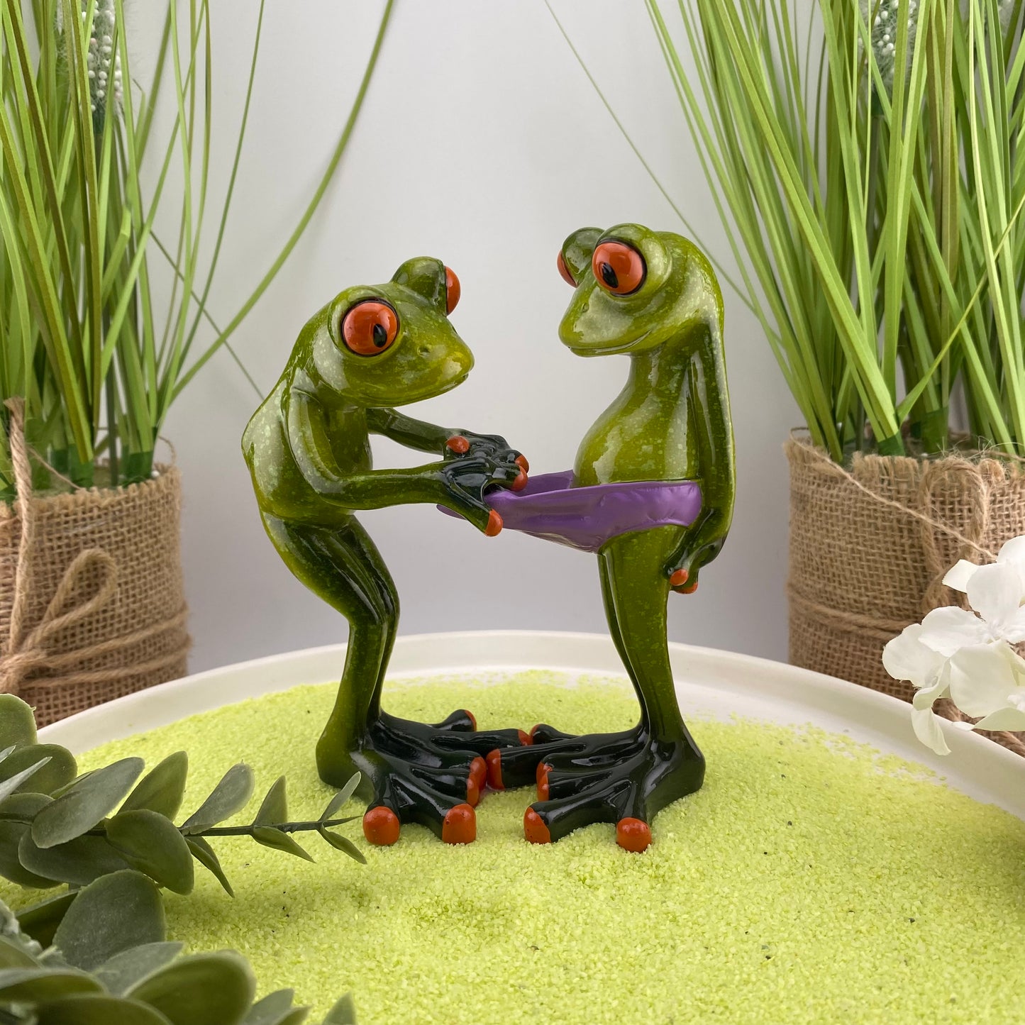 Eine Dekofigur im Ambiente, die ein Froschpaar abbildet. Der eine Frosch zieht frech an der Hose des anderen Frosches und schaut hinein.