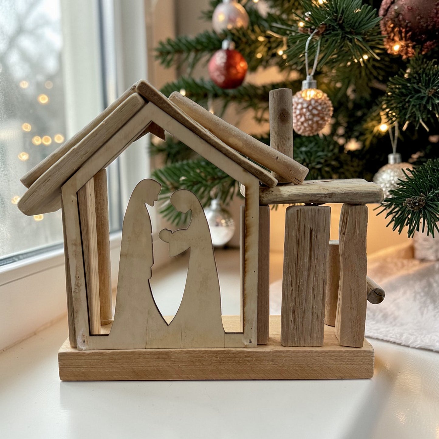 Schlichte Krippe aus Holz mit minimalistischer Silhouetten-Darstellung, seitlich aufgenommen. Platziert auf einer Fensterbank mit geschmücktem Weihnachtsbaum und warmem Licht im Hintergrund.