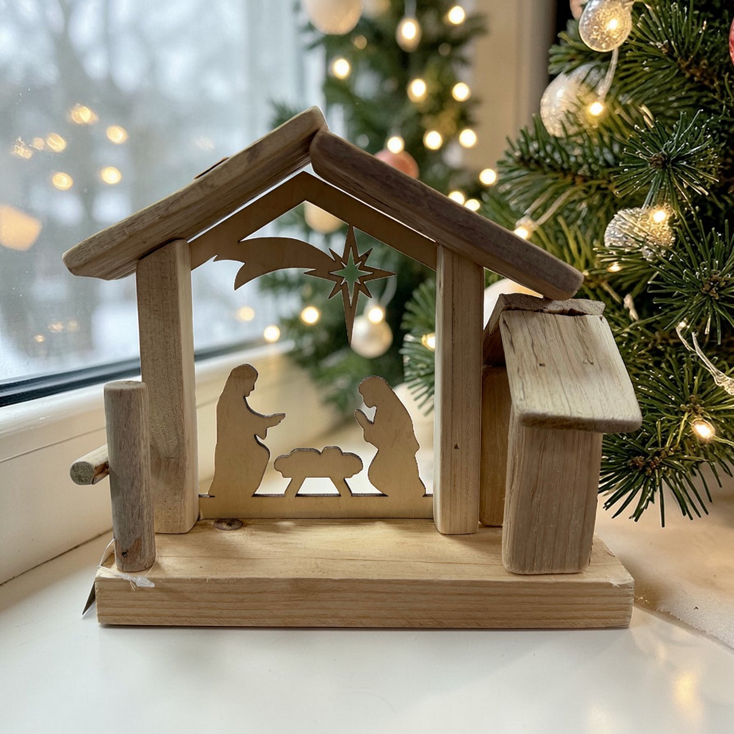 Schlichte Krippe aus Holz mit ausgeschnittener Szene von Maria, Josef und dem Kind, ergänzt durch einen Stern über dem Stall. Platziert vor einem geschmückten Weihnachtsbaum mit warmem Licht im Hintergrund.