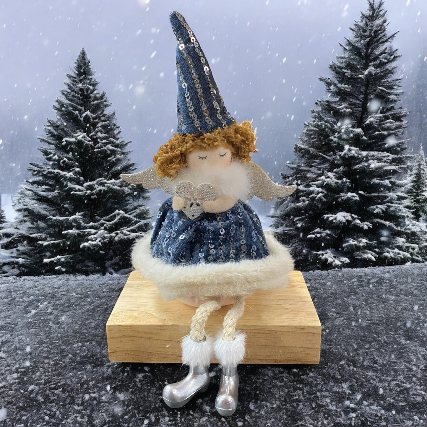Sitzende Stoff-Engelfigur mit blauen Paillettenkleid, weißen Flügeln und Mütze auf einem Holzblock, im Hintergrund verschneite Tannenbäume.