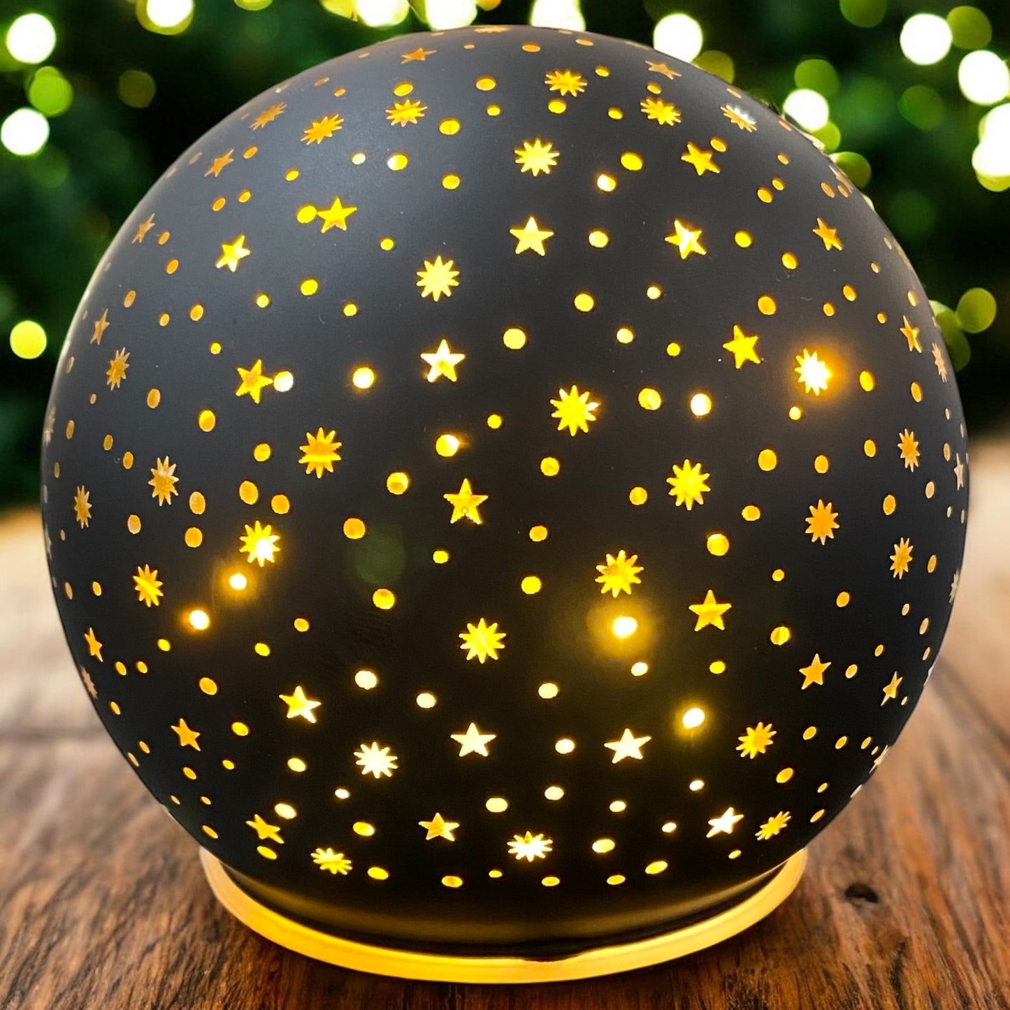 Leuchtende schwarze Deko-Kugel mit LED-Beleuchtung und Sternenmuster, dekorativ auf Holztisch vor Lichterhintergrund platziert.