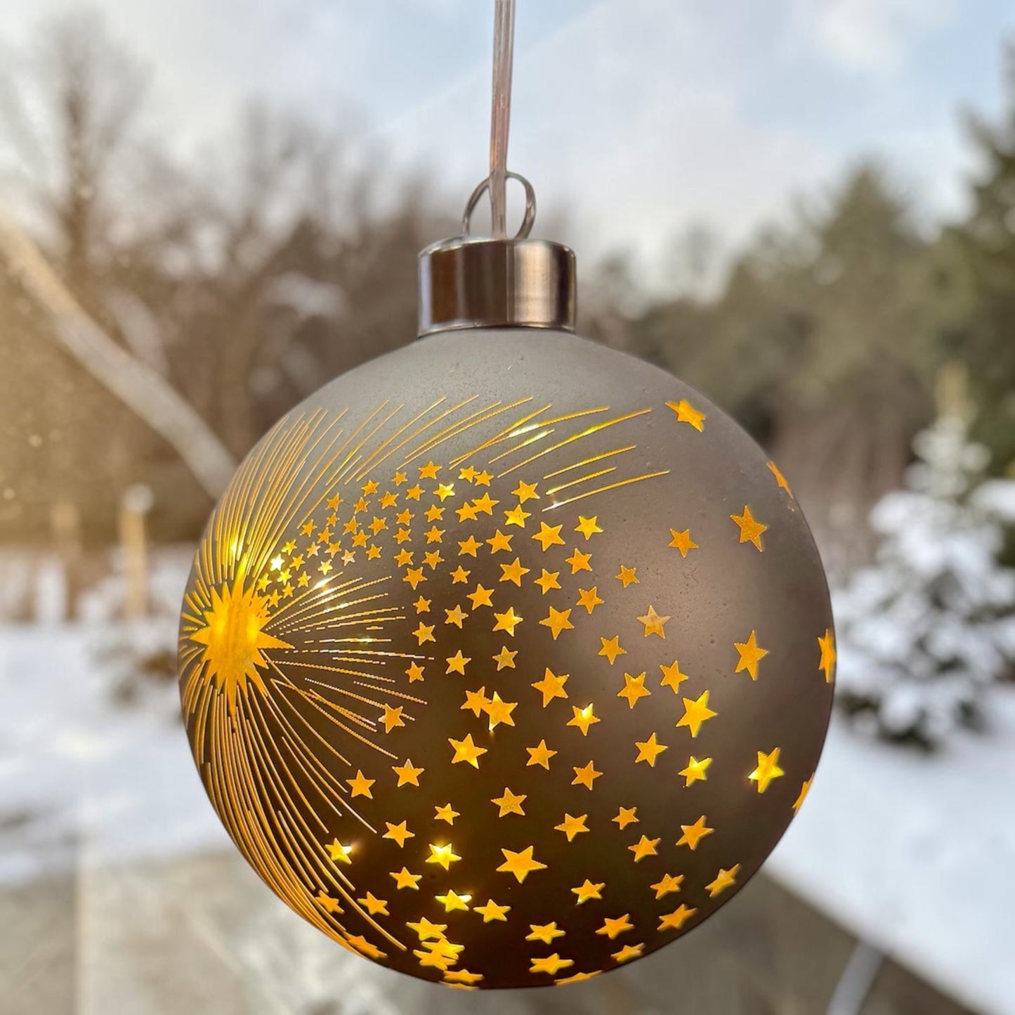 Eine taupefarbene LED-Dekokugel mit Sternschnuppenmotiv hängt vor einer Fensterfläche mit unscharfem Außenbereich.