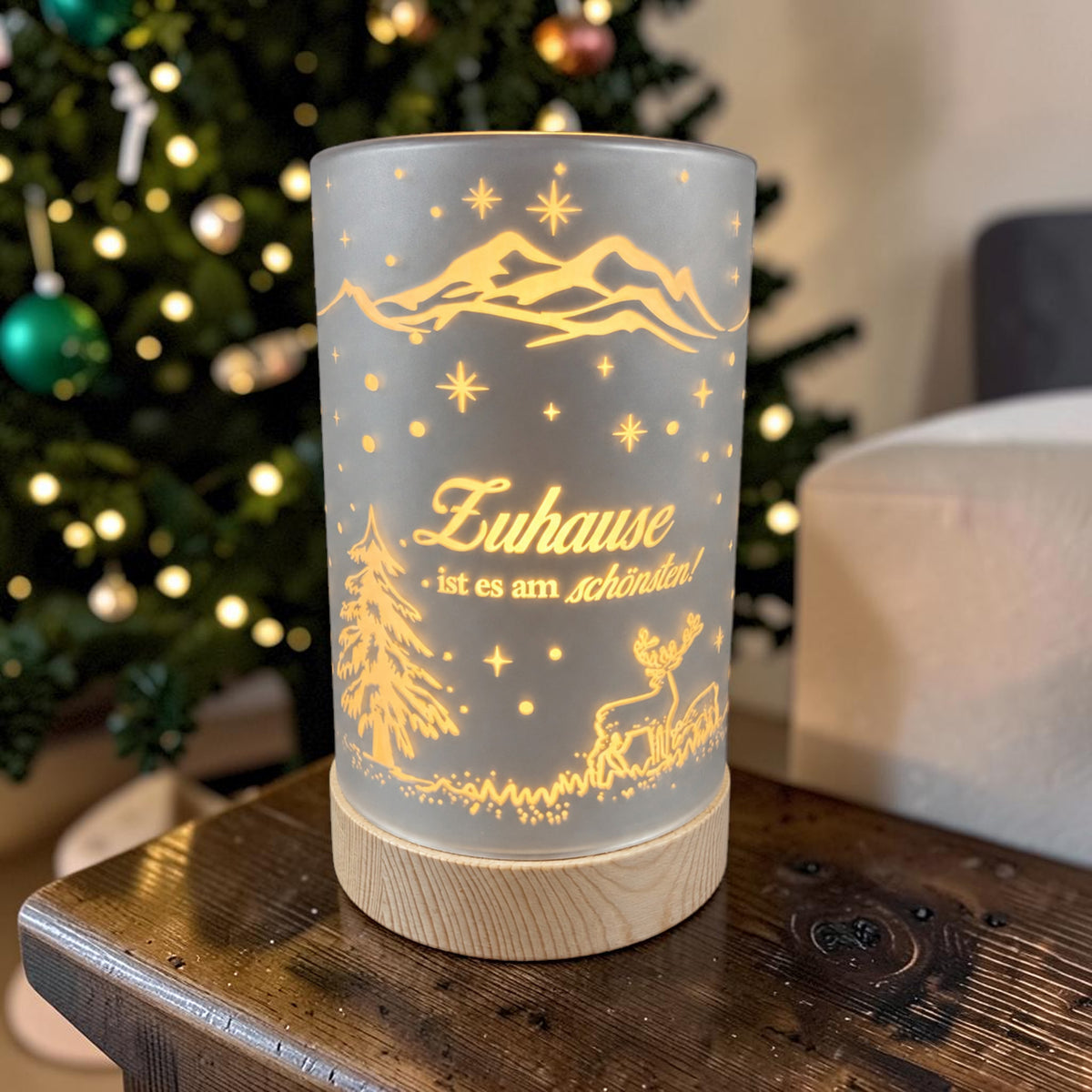 Kleine silberne Formano LED Säule „Zuhause ist es am schönsten!“ mit Holzsockel und warm leuchtendem Wintermotiv vor einem Weihnachtsbaum.