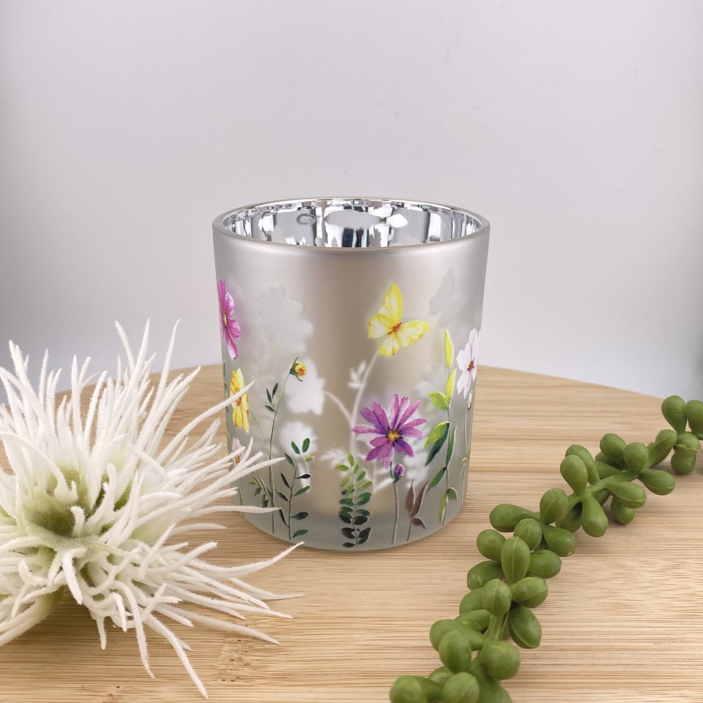 Teelichtglas in silber mit bunten Blumen und Schmetterlingen im Ambiente platziert