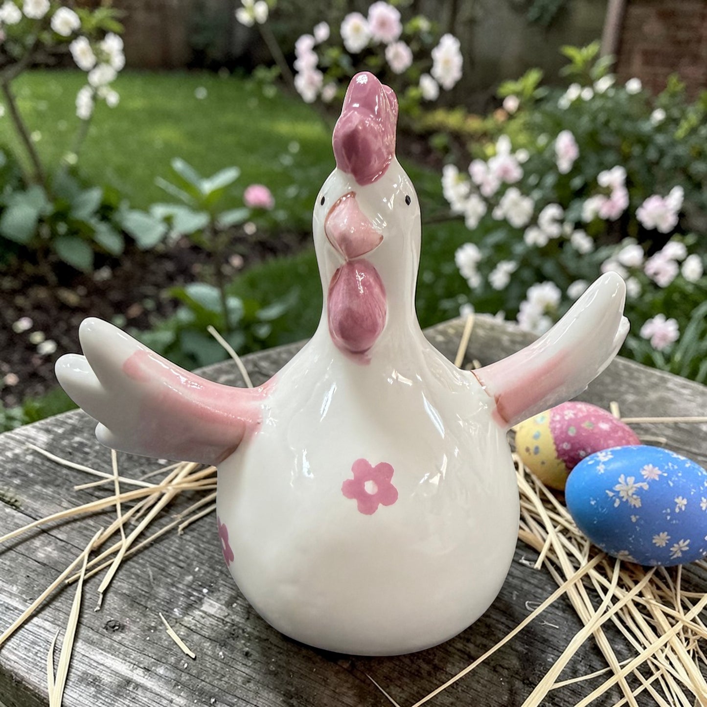 Frühlingshafte Osterdekoration mit Keramik-Huhn in Cremeweiß und Rosa, arrangiert auf einer rustikalen Holzoberfläche. Umgeben von Stroh, bunten Ostereiern und blühenden Pflanzen entsteht eine natürliche, lebendige Gartenatmosphäre.