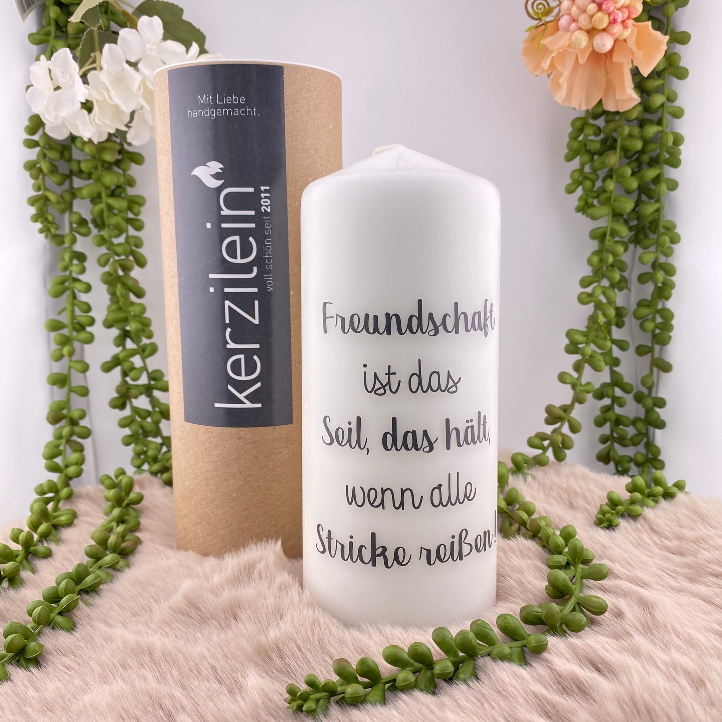 Eine große, weiße Kerze mit schwarzem Schriftzug "Freundschaft ist das Seil, das hält, wenn alle Stricke reißen". Die Dekokerze ist 18,5cm groß und im Ambiente platziert.