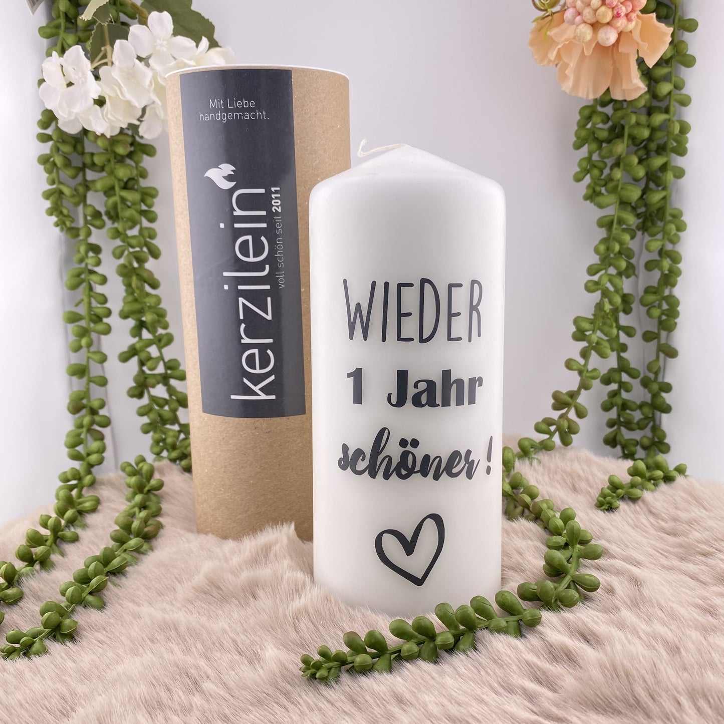 Eine große, weiße Kerze mit schwarzem Schriftzug "Wieder 1 Jahr schöner". Die Dekokerze ist 18,5cm groß und im Ambiente platziert.