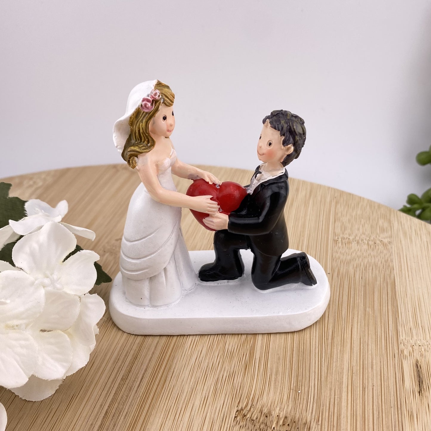 Hochzeit Dekofigur Aufsteller auf weißem Sockel im Ambiente bildet Brautpaar. Der Mann in schwarzem Anzug kniet vor Frau im Brautkleid und überreicht ihr ein großes Rotes Herz als Symbol seiner Liebe.