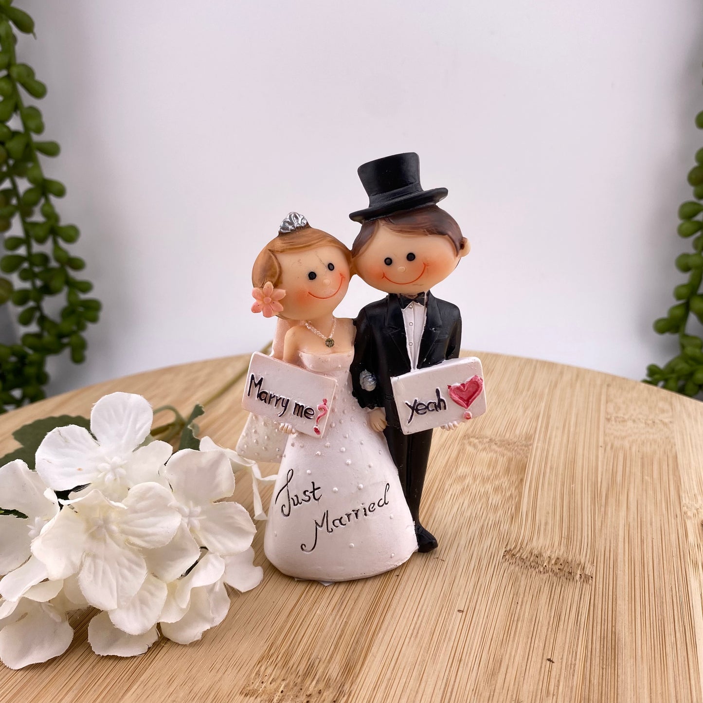 Hochzeit Dekofigur Tortenaufsteller im Ambiente bildet Brautpaar Mann & Frau ab. Das Ehepaar hält sich an der Hand. Die Frau hält ein Schild mit "Marry Me?" und der Mann mit "Yeah". Auf dem weißen Brautkleid steht Just Married.