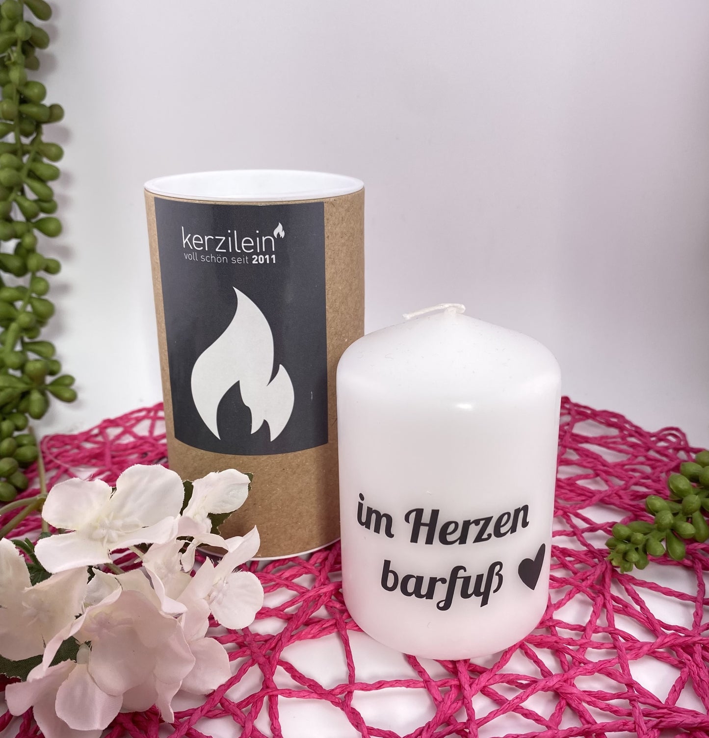 Eine kleine, weiße Kerze mit schwarzem Schriftzug "Im Herzen barfuß". Die Dekokerze ist 8cm groß und im Ambiente platziert.