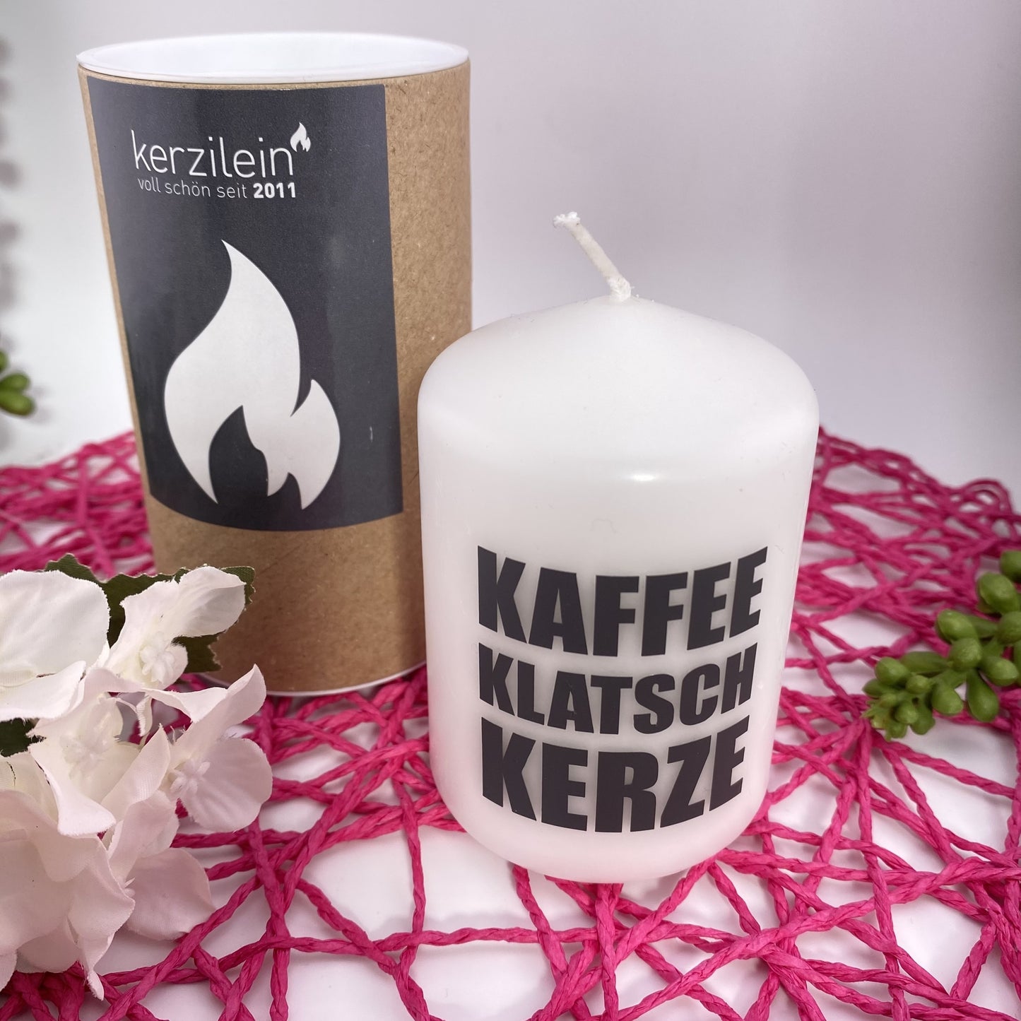 Eine kleine, weiße Kerze mit schwarzem Schriftzug "Kaffeeklatsch". Die Dekokerze ist 8cm groß und im Ambiente platziert.