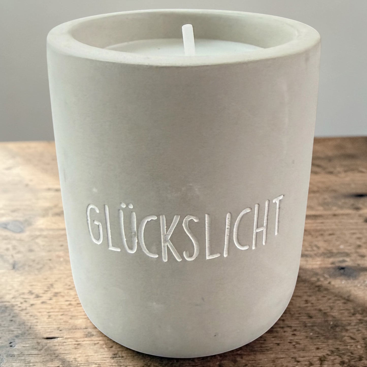 Graue Betonkerze mit eingraviertem Schriftzug „GLÜCKSLICHT“ auf einer Holzoberfläche.