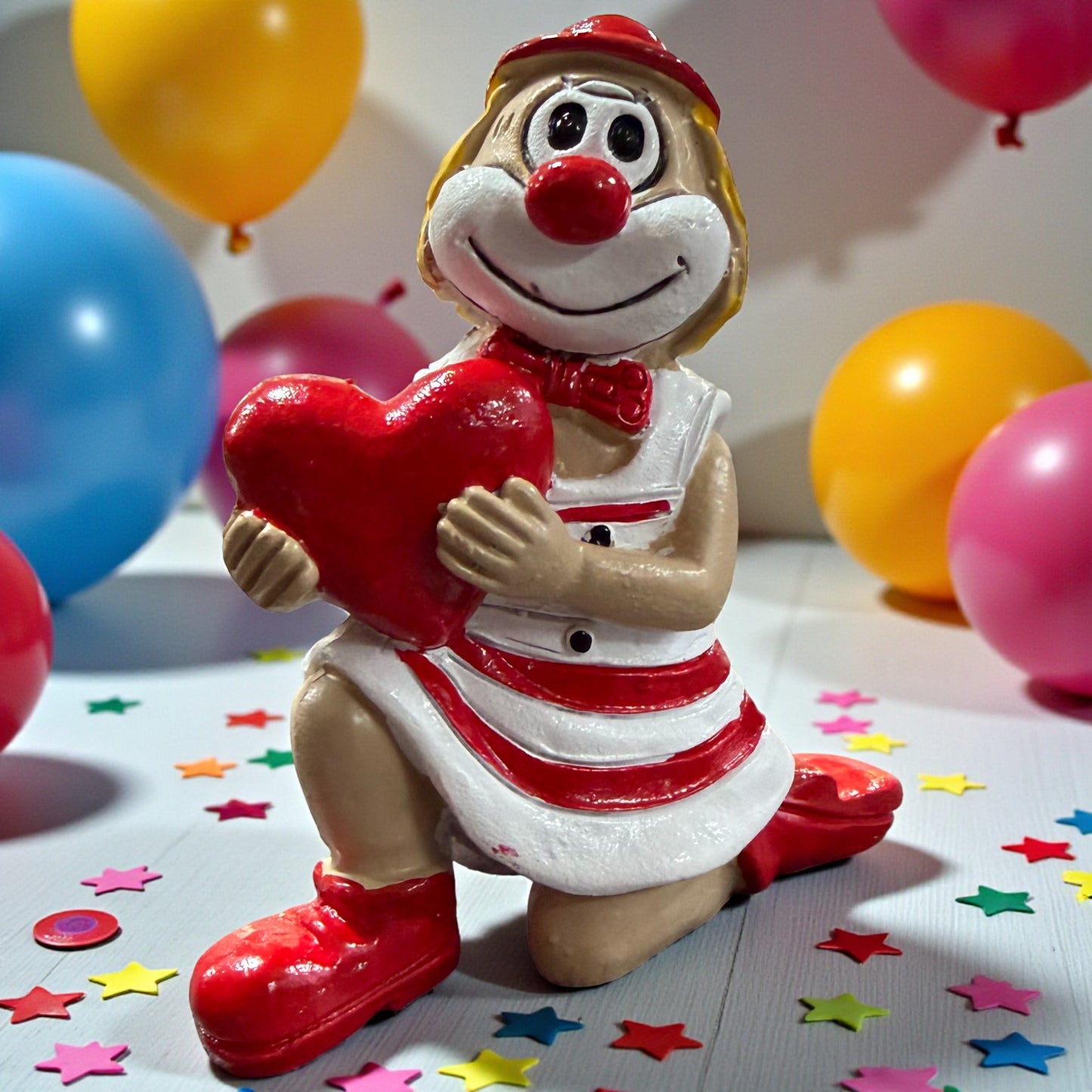 Clown-Figur mit roter Nase in rot-weißem Outfit, kniend dargestellt und ein rotes Herz haltend, auf einer Tischfläche mit Konfetti, im Hintergrund farbige Luftballons.