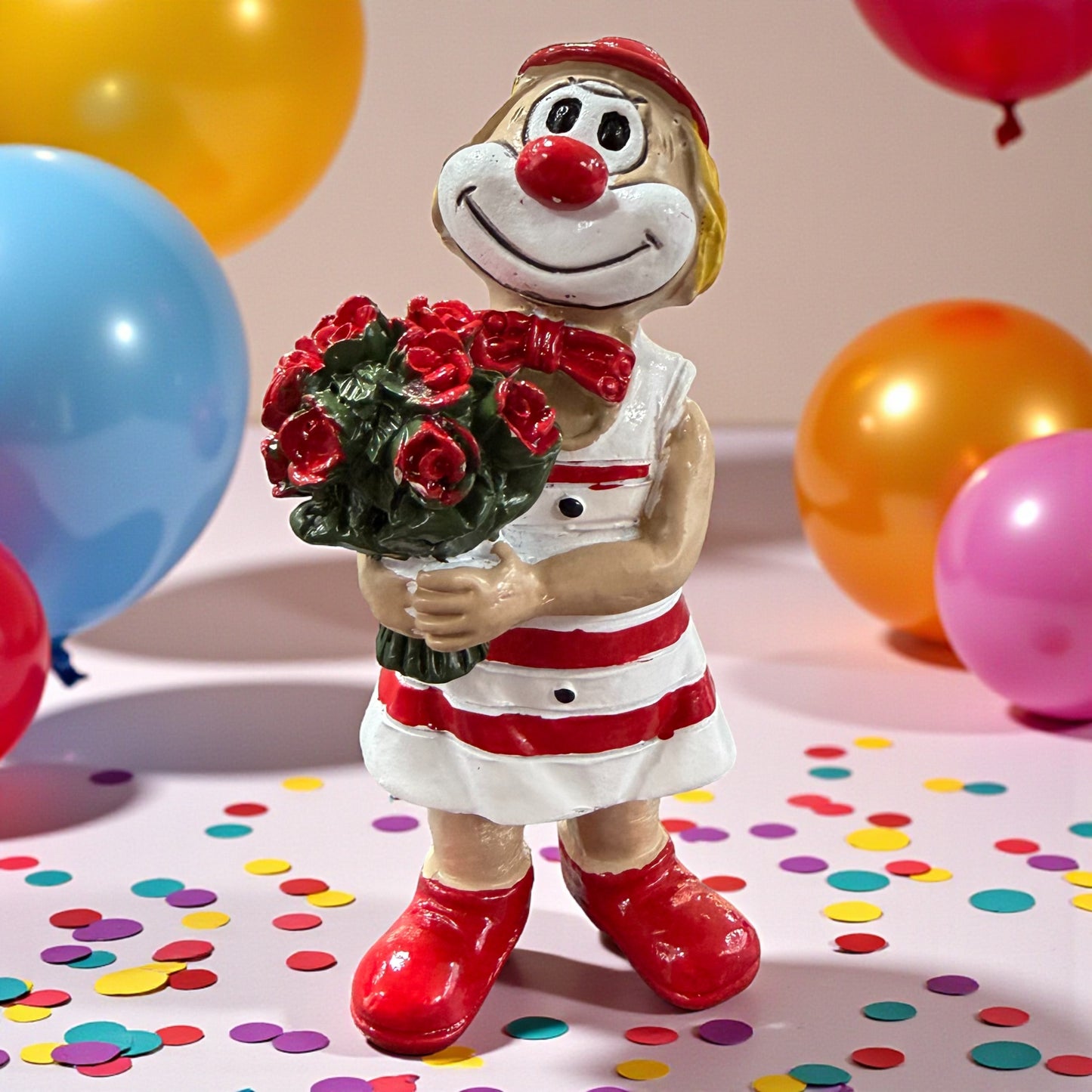 Clown-Figur mit roter Nase in rot-weißem Outfit, hält einen Blumenstrauß mit roten Rosen, stehend auf einer Tischfläche mit Konfetti, im Hintergrund farbige Luftballons.