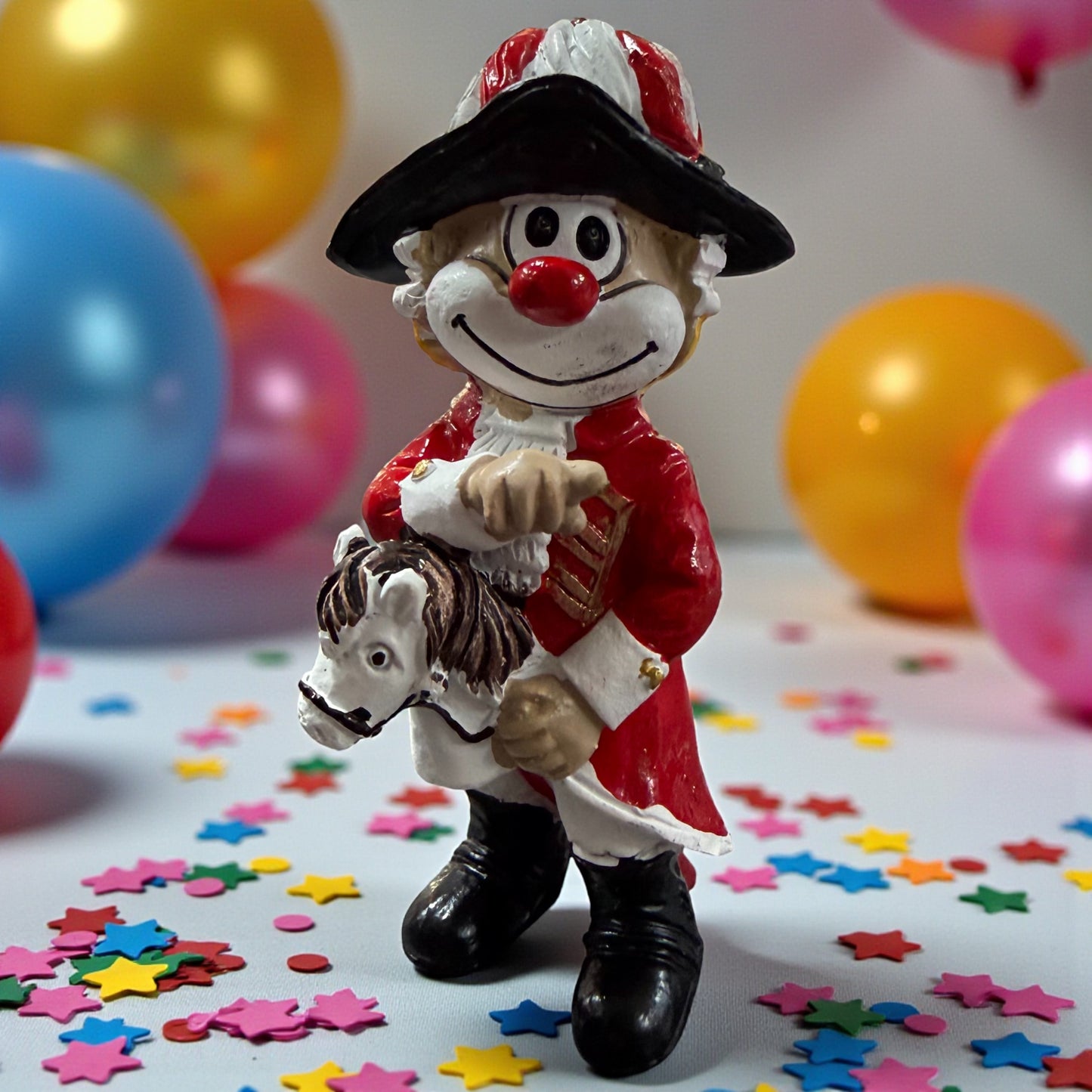 Clown-Figur mit roter Nase in roter Uniform und schwarzem Hut, hält ein kleines Pferd vor dem Körper, stehend auf einer Tischfläche mit Konfetti, im Hintergrund farbige Luftballons.