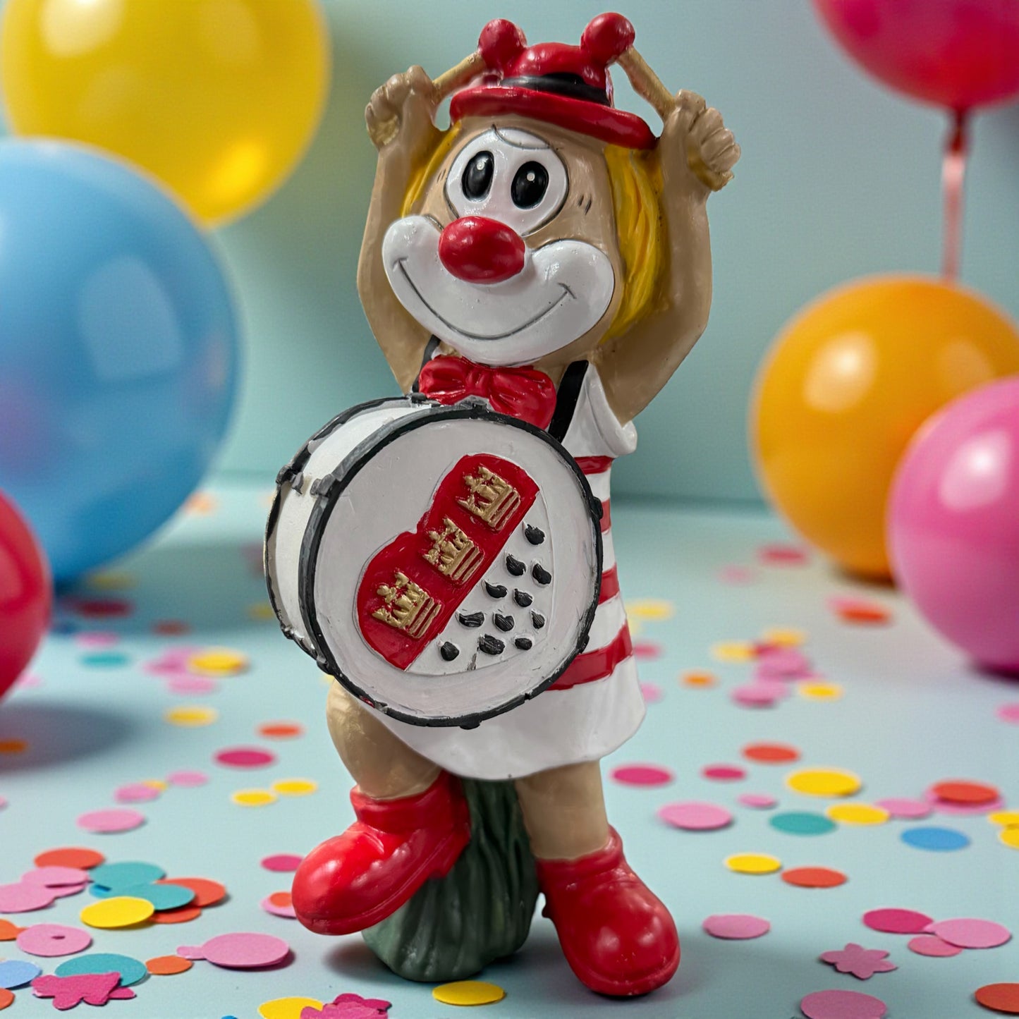 Große Clown-Dekofigur mit roter Nase in rot-weißem Outfit, ca. 20 cm groß, stehend dargestellt und eine Trommel mit kölschem Herzmotiv haltend, auf einer Tischfläche mit Konfetti, im Hintergrund bunte Luftballons.