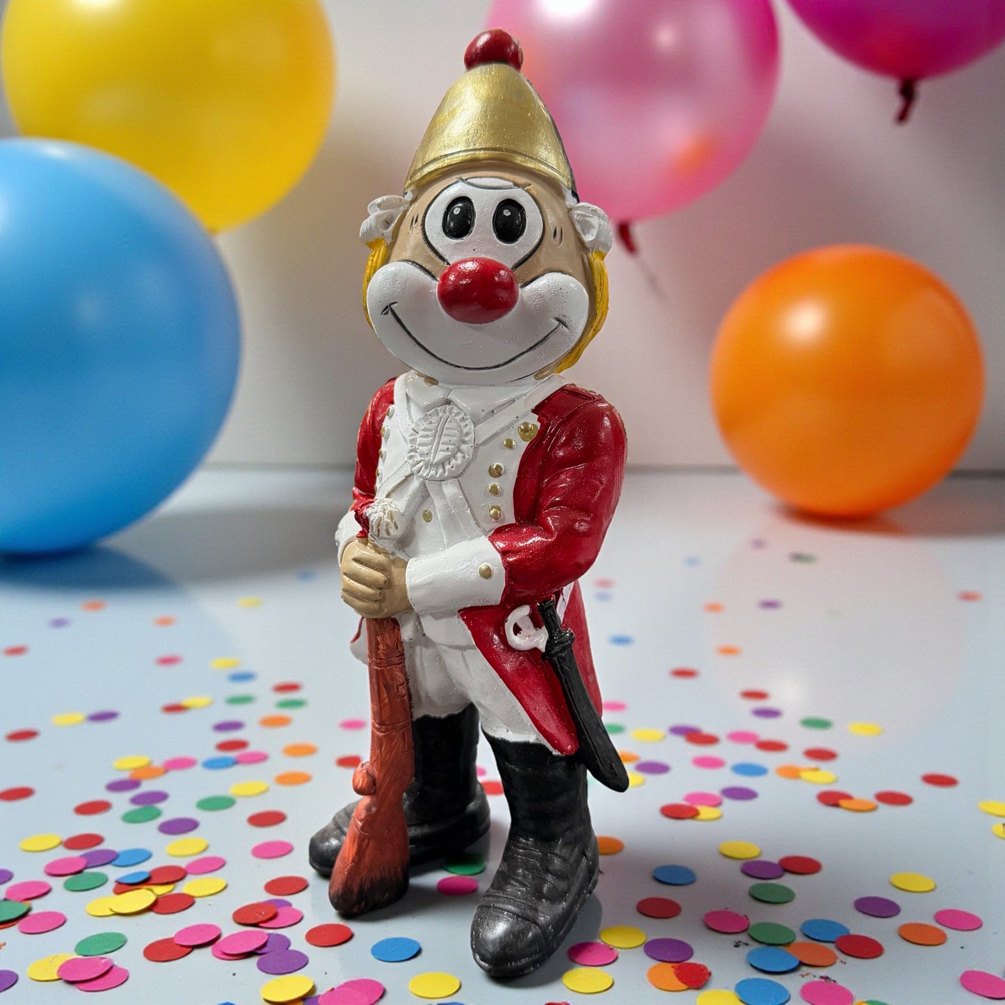 Große Clown-Dekofigur mit roter Nase in rot-weißer Fußkorps-Uniform und goldfarbenem Helm, ca. 20 cm groß, stehend dargestellt und ein Gewehr haltend, auf einer Tischfläche mit Konfetti, im Hintergrund bunte Luftballons.