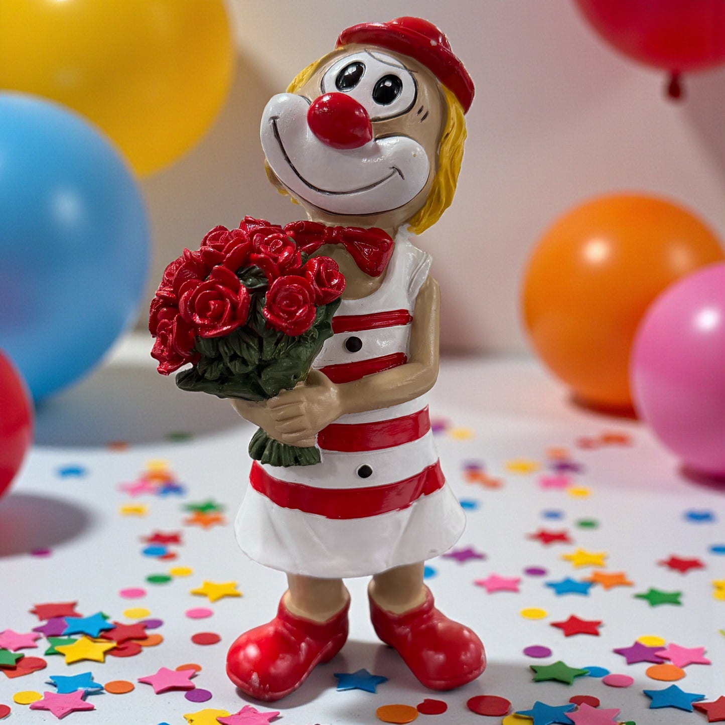 Große Clown-Dekofigur mit roter Nase in rot-weißem Outfit, ca. 20 cm groß, stehend dargestellt und einen Strauß roter Rosen haltend, auf einer Tischfläche mit Konfetti, im Hintergrund bunte Luftballons.