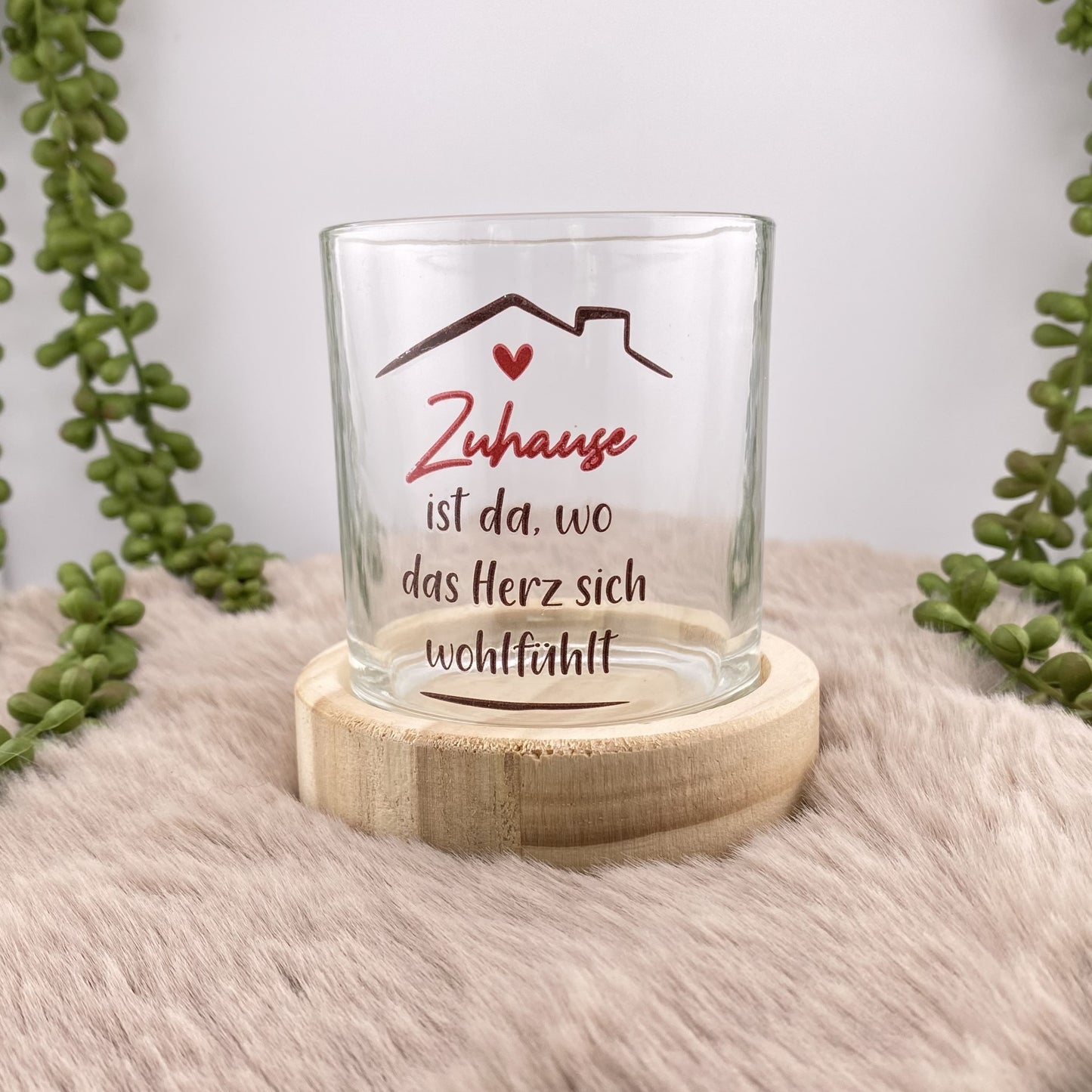 Ein Teelichthalter mit rundem Holzsockel und Glas mit rotem Schriftzug "Zuhause ist da, wo das Herz sich wohlfühlt" ist im Ambiente platziert