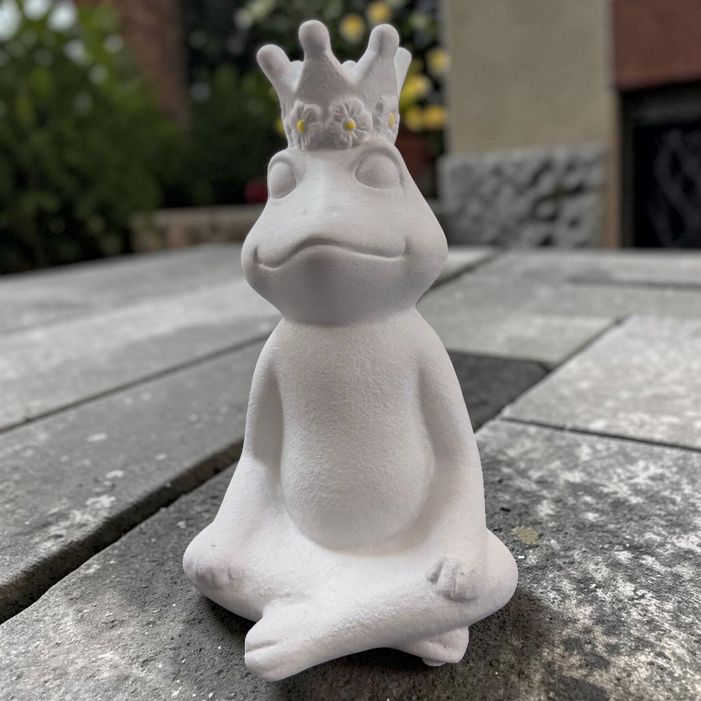 Großer sitzender Frosch in matter Weiß-Optik mit dekorativer Krone auf rustikalem Gartentisch.