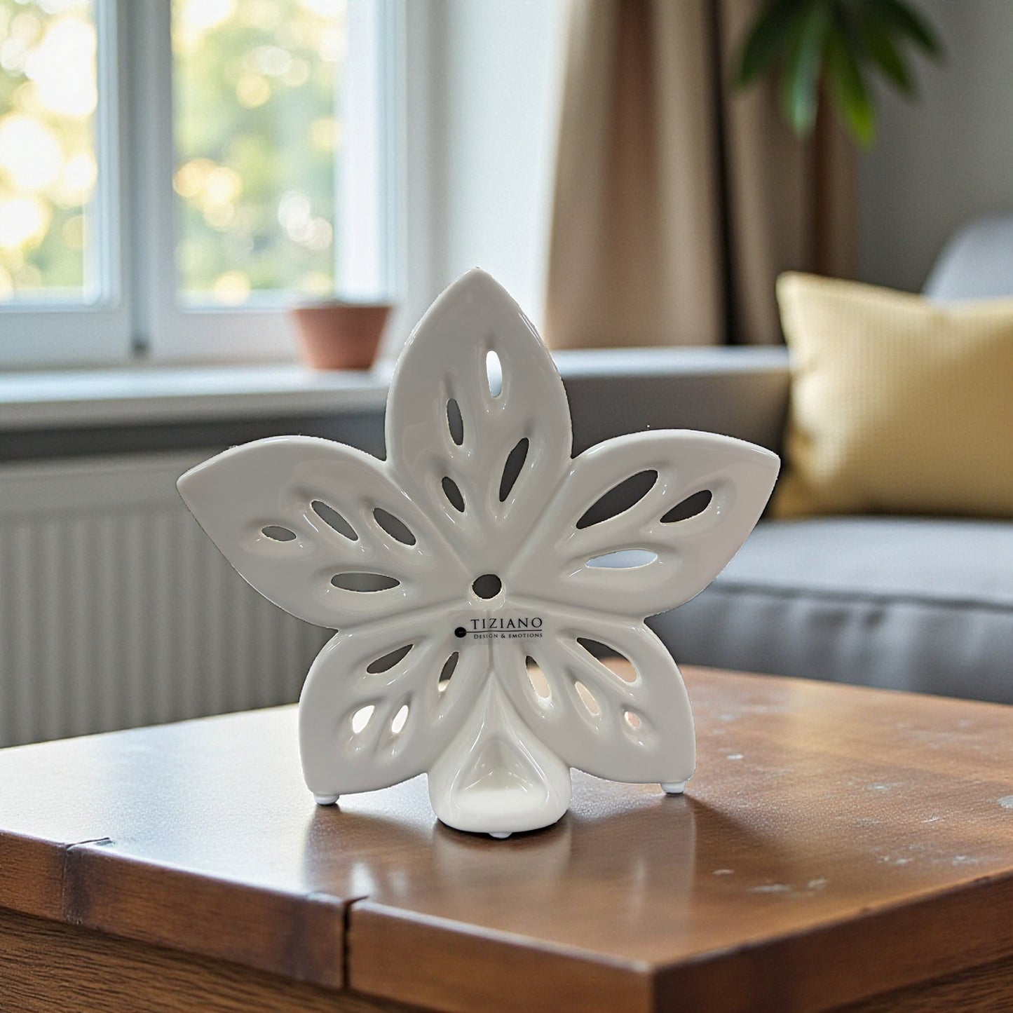 Eine creme-weiße Keramikblume mit Lochmuster, aufgestellt auf einem Holztisch vor einem Fenster mit Heizkörper und Sitzmöbeln im Hintergrund.