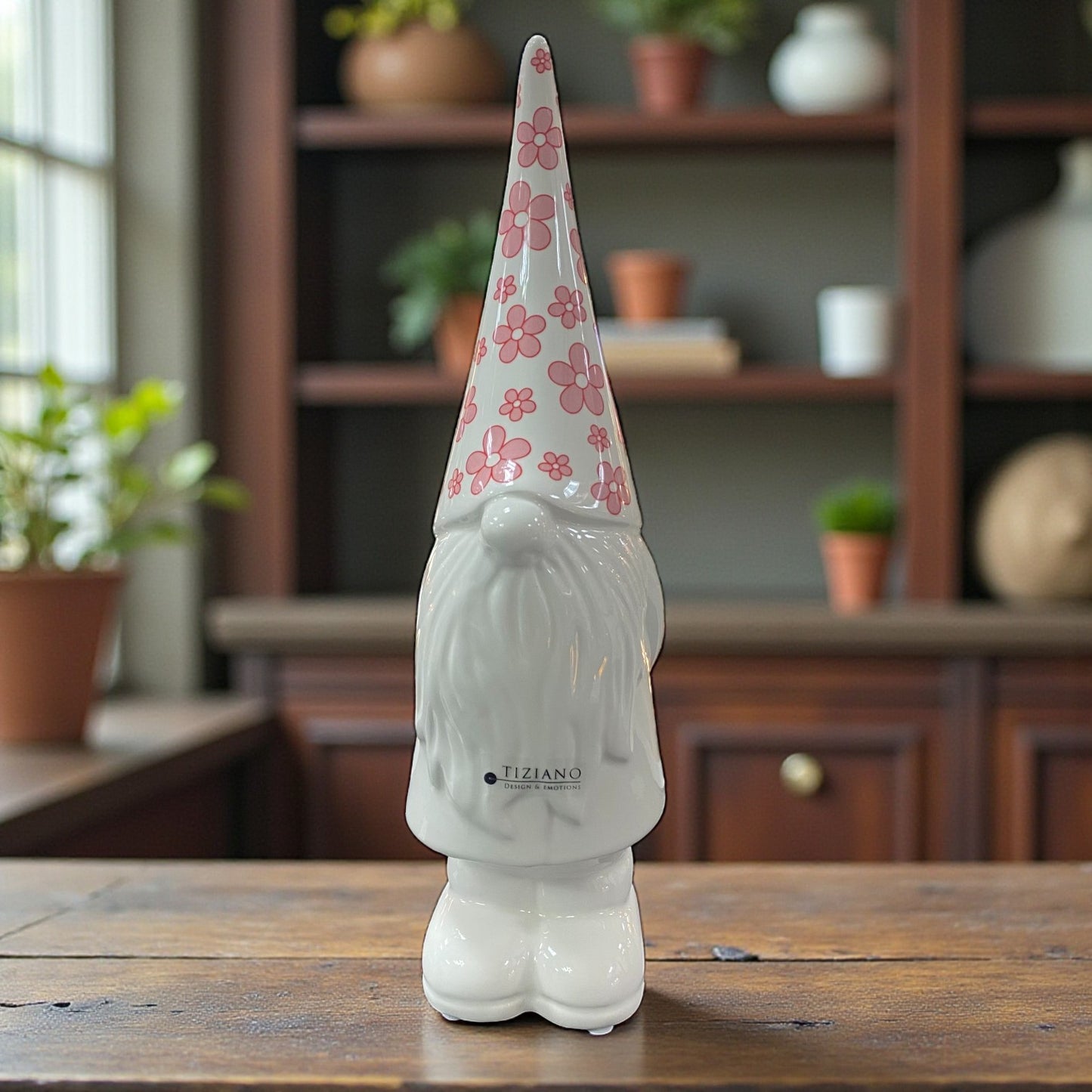 Weiße Keramik-Zwergfigur mit langer Zipfelmütze im rosa Blumenmuster, frontal stehend auf Holztisch in Wohnraum mit Regal im Hintergrund