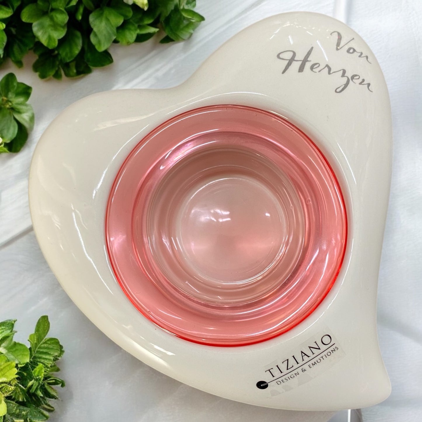 Ein creme-weißer Keramik-Teelichthalter in Herzform von Tiziano, mit integriertem Glaseinsatz in Pink und Schriftzug „Von Herzen“ auf der Oberfläche, im Ambiente