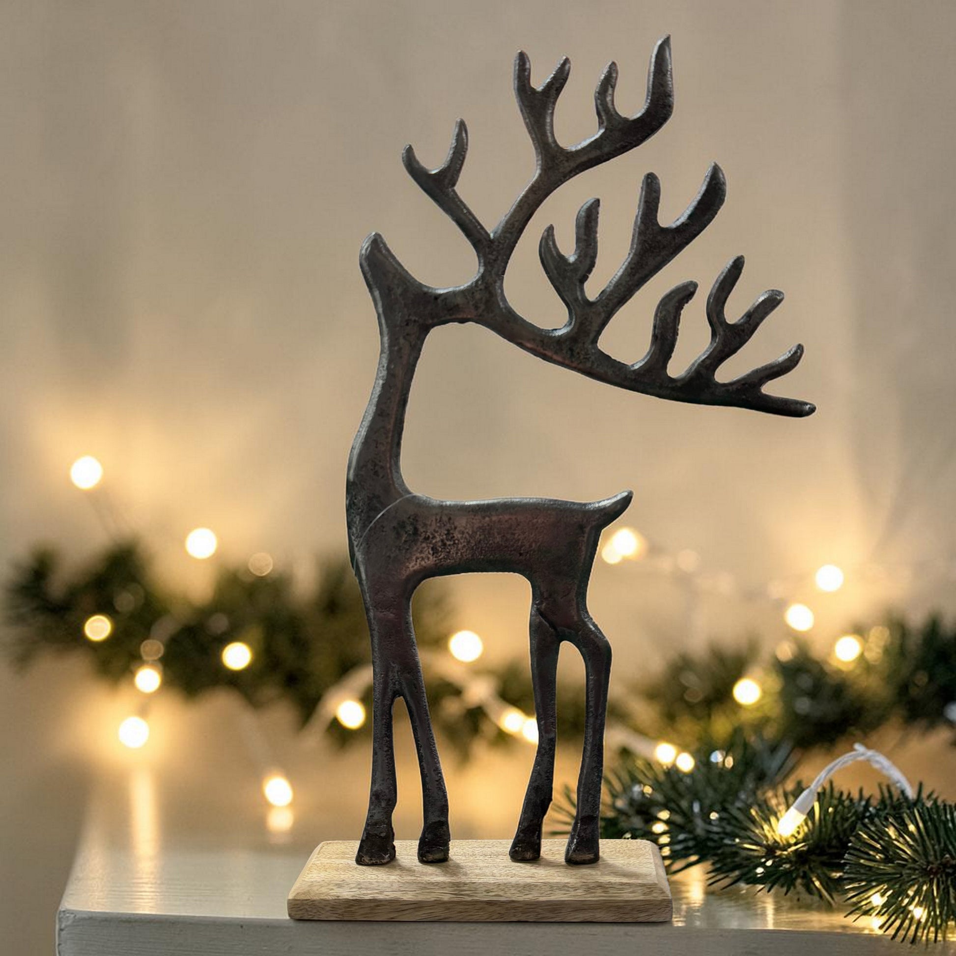 Eine stilisierte Hirschfigur aus dunklem Material steht auf einem Holzsockel vor einer Fläche mit Lichterkette im Hintergrund.
