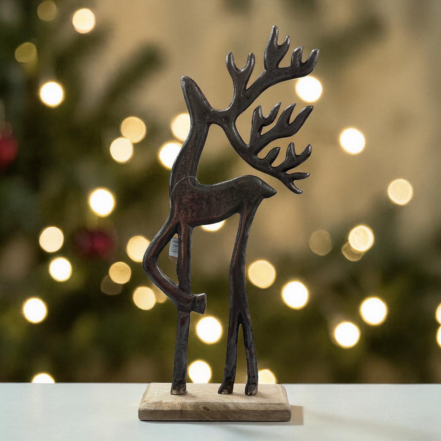 Eine stilisierte Hirschfigur aus dunklem Material steht auf einem Holzsockel vor einem unscharfen Hintergrund mit warmen Lichtpunkten.