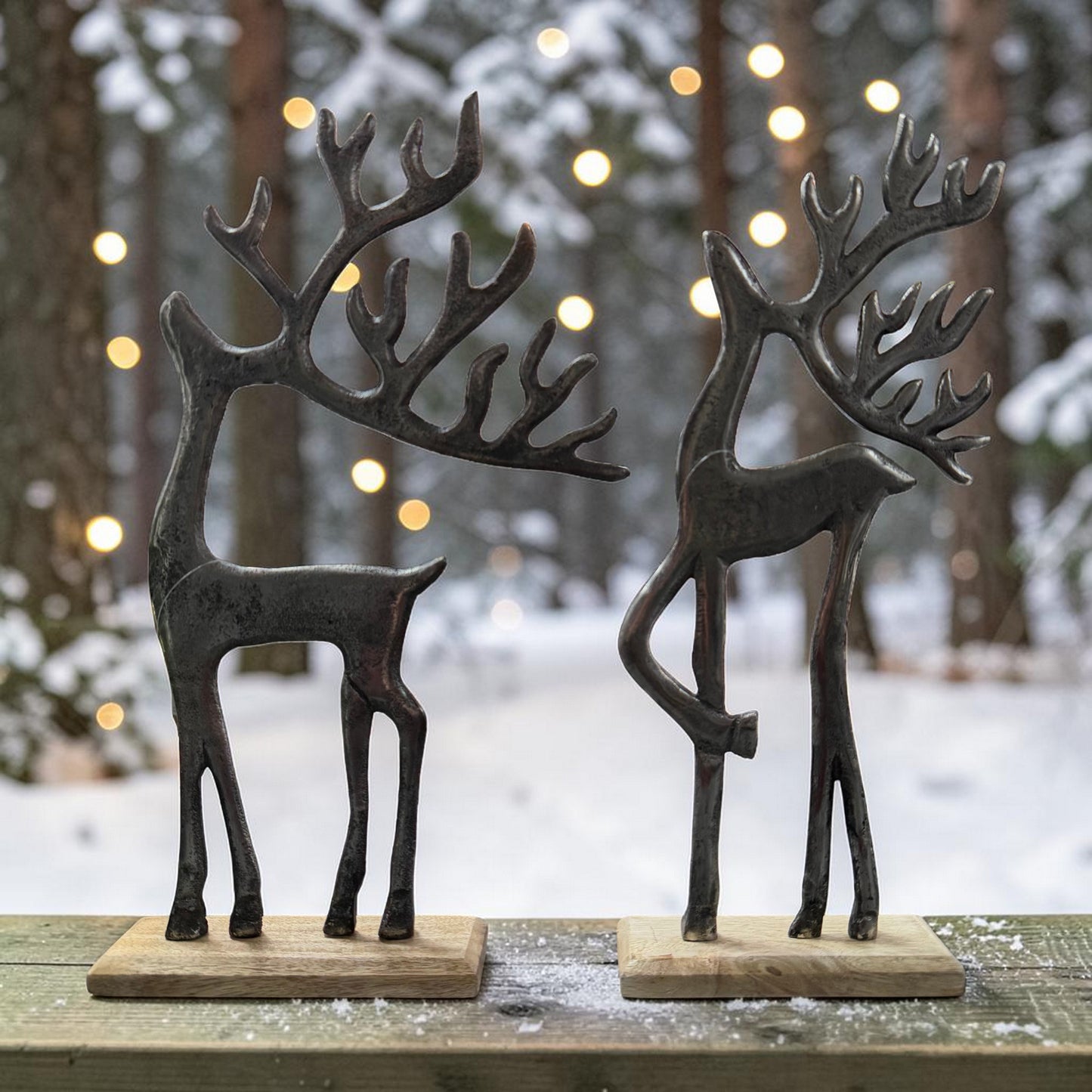 Zwei stilisierte Hirschfiguren aus dunklem Material stehen auf einer Holzfläche vor einem unscharfen winterlichen Hintergrund mit Lichtern.