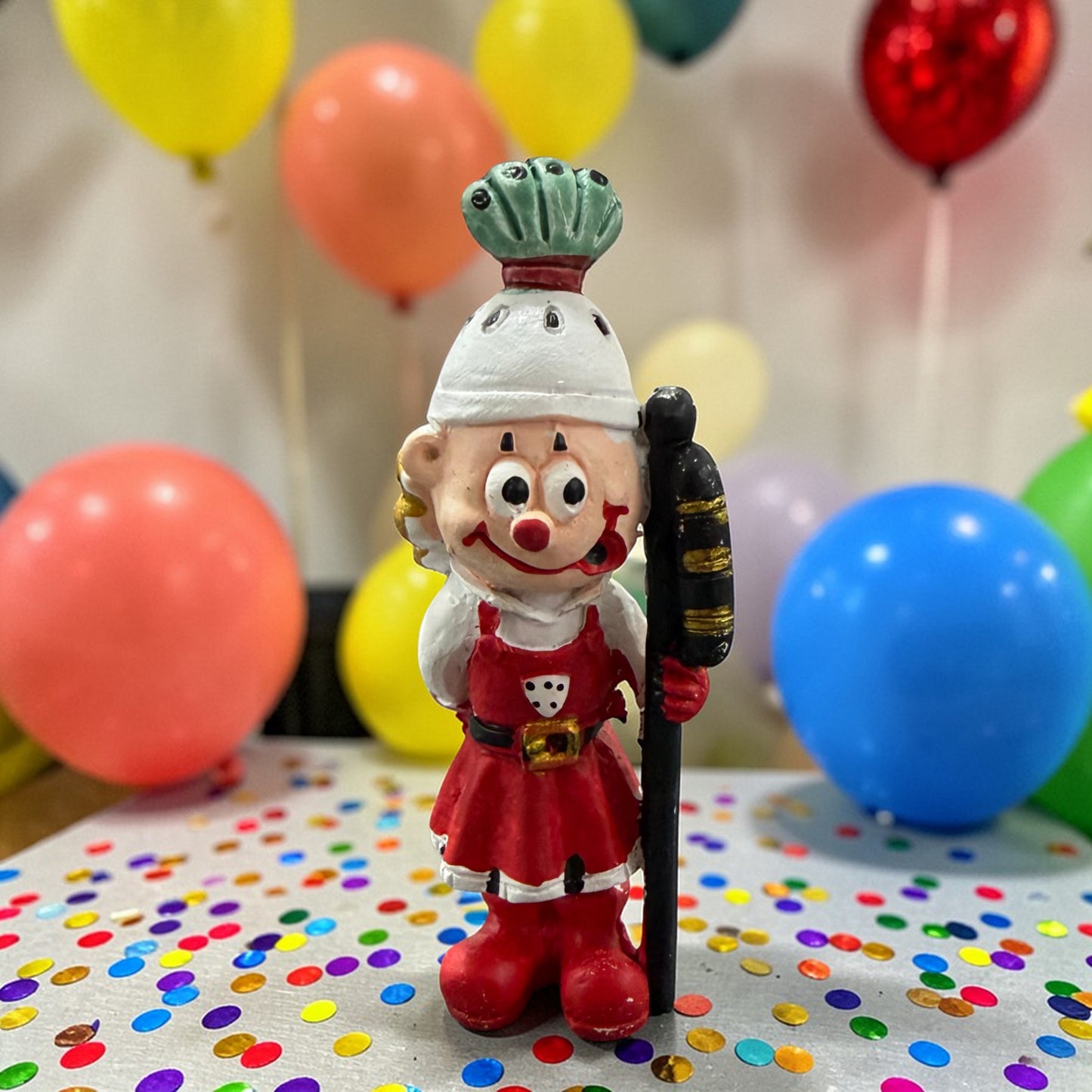 Clown-Dekofigur Bauer mit Mütze und Stab vor Luftballons und Konfetti.