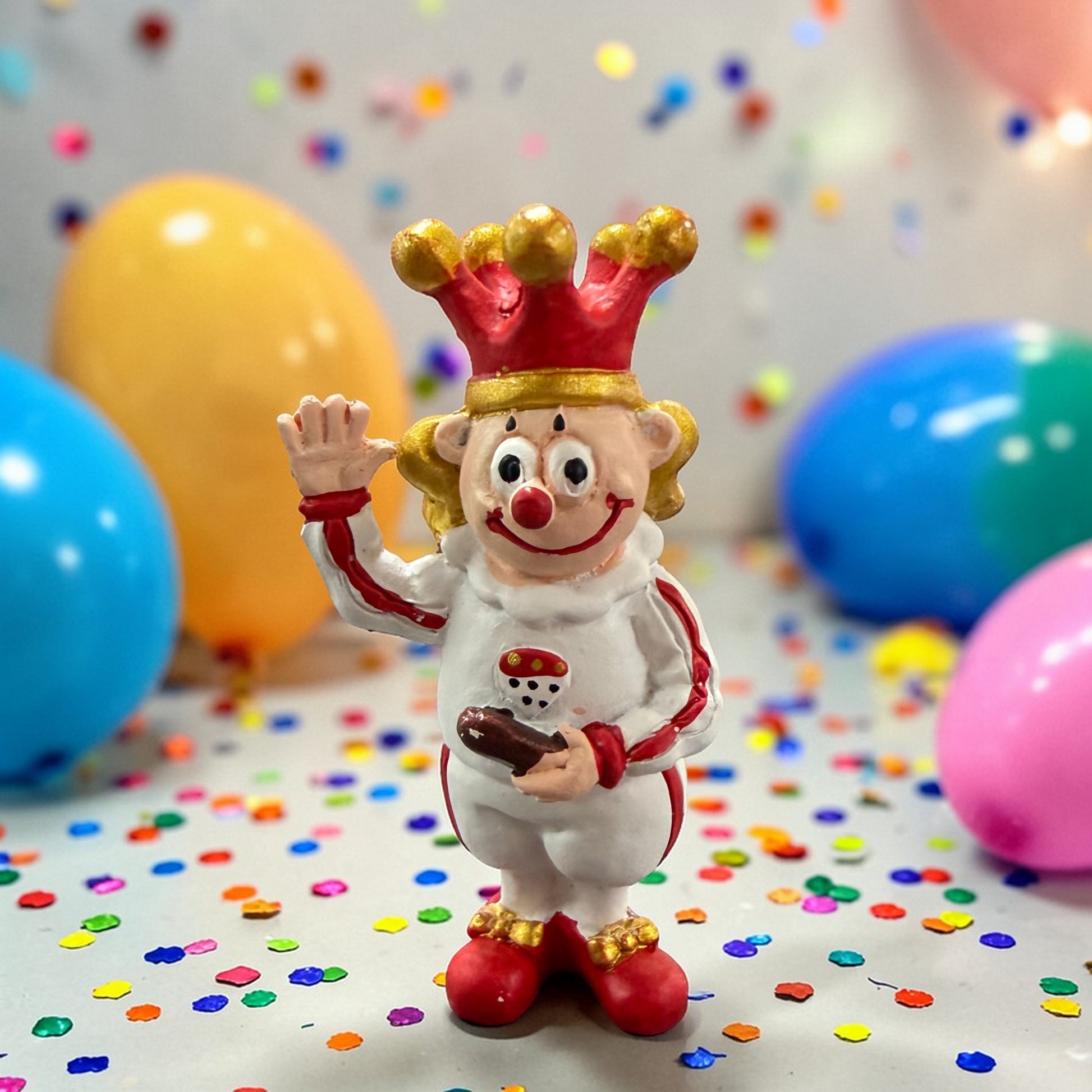 Clown-Dekofigur Prinz mit Krone und Zepter vor Luftballons und Konfetti.