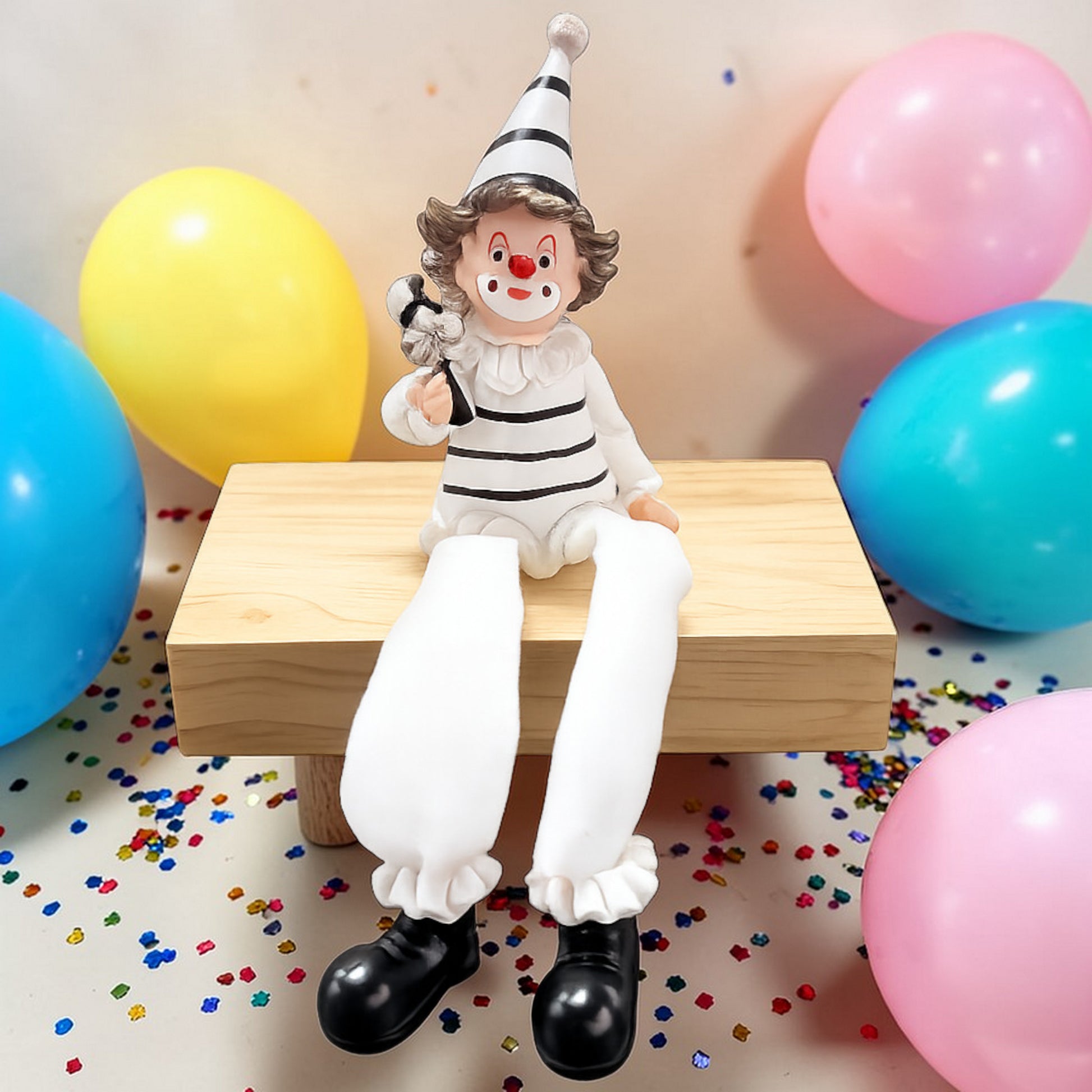 Clown-Kantenhocker in Schwarz-Weiß mit Mütze auf Holzblock vor Luftballons und Konfetti.