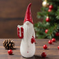 Ein Keramik-Wichtel mit roter Mütze hält ein Geschenk in der Hand und ist stimmungsvoll vor einem Weihnachtsbaum arrangiert.