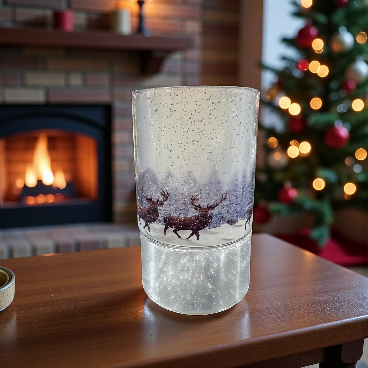 Ein zylindrisches Glaslicht mit winterlichem Hirschmotiv steht auf einer Holzfläche vor einem Kamin und einem geschmückten Weihnachtsbaum.