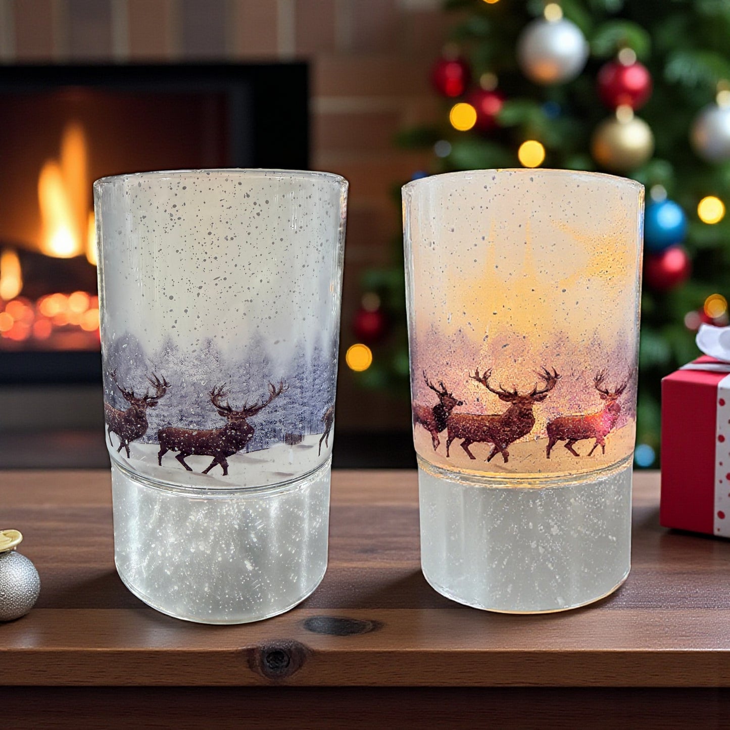 Zwei zylindrische Glaslichter mit winterlichem Hirschmotiv stehen auf einer Holzfläche vor einem Kamin und einem geschmückten Weihnachtsbaum.
