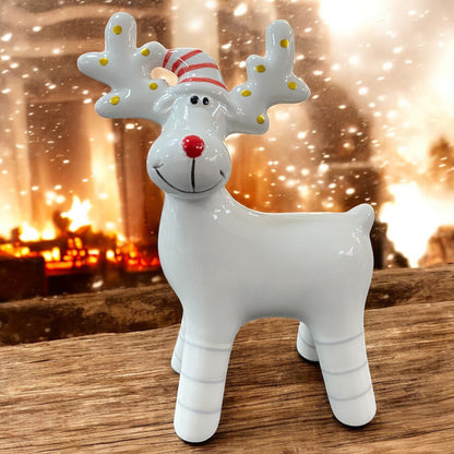 Kleines weißes Porzellan-Rentier mit goldenen Punkten und roter gestreifter Mütze, vor Kaminfeuer und Schneeflocken dekoriert.